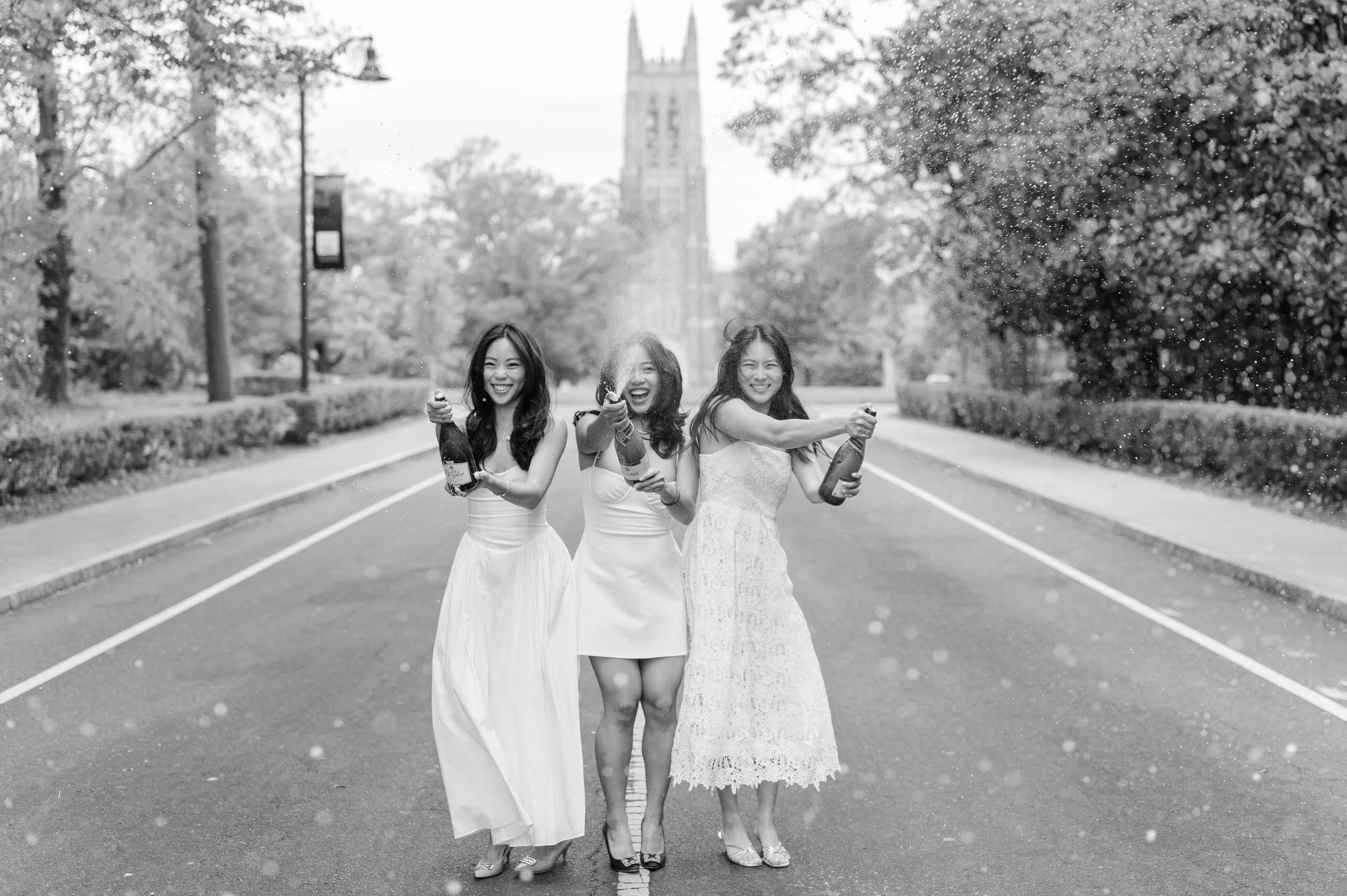 Three women in white dresses standing on a street, celebrating with champagne bottles, spraying champagne, smiling, with a cityscape and clock tower in the background.