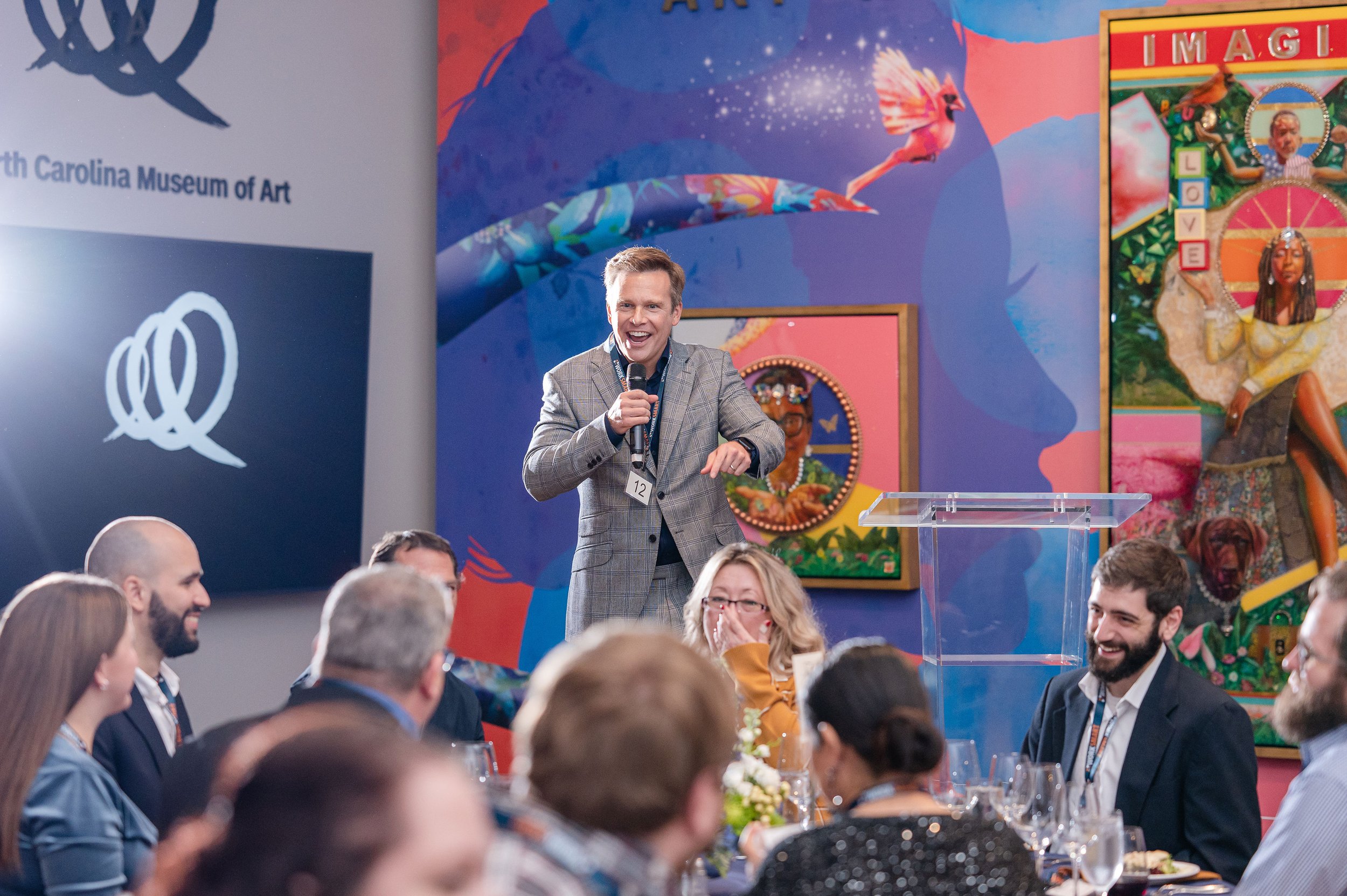 A man in a plaid suit jacket stands at a podium holding a microphone, speaking to an audience at a conference or event. The background features colorful wall art and a large banner with the logo of the North Carolina Museum of Art. People seated at round tables with wine glasses are listening and smiling.