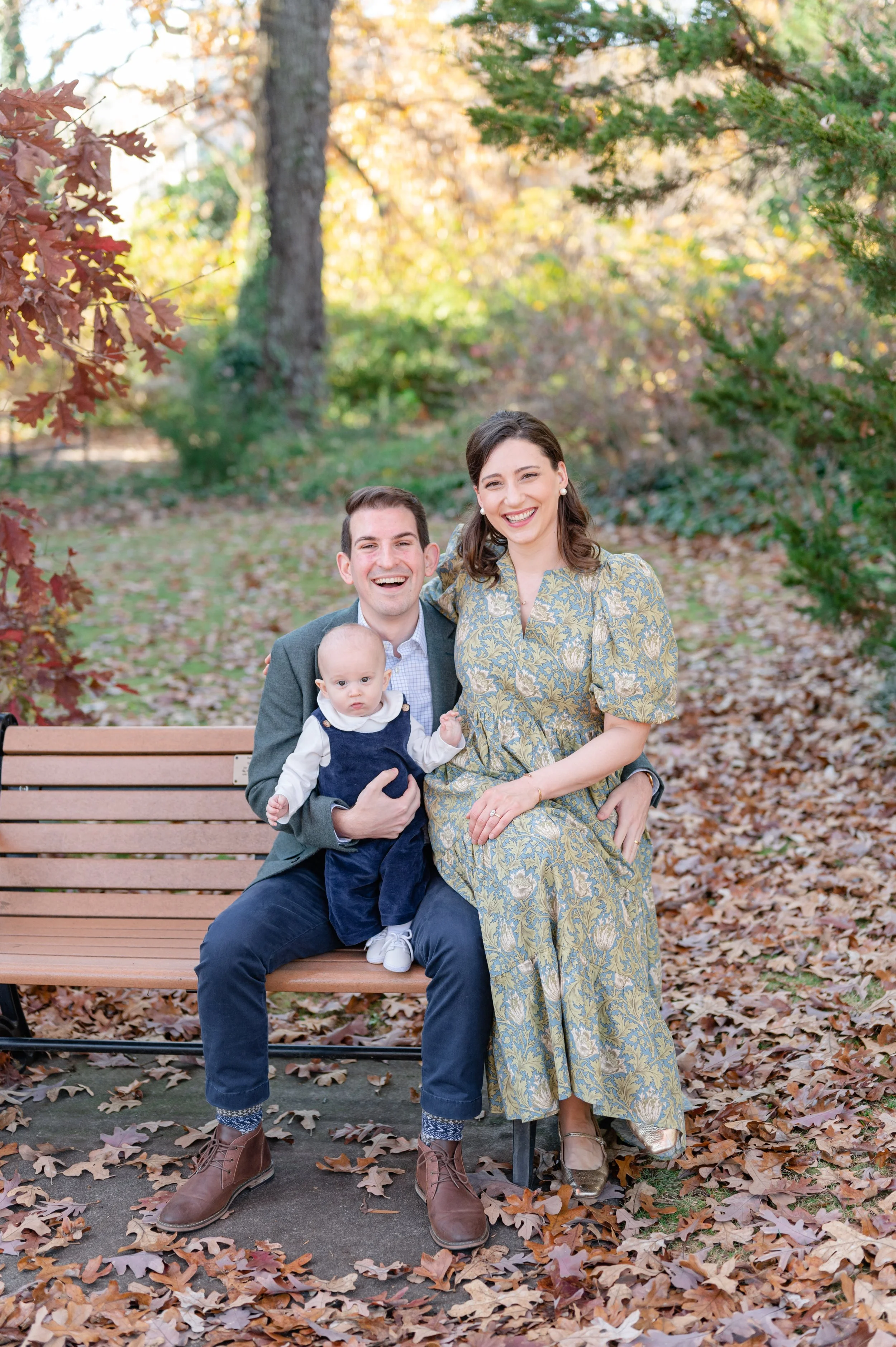A family of three sitting on a park bench surrounded by fallen autumn leaves, with trees and foliage in the background. The man holds a baby, and the woman sits beside them, all smiling.