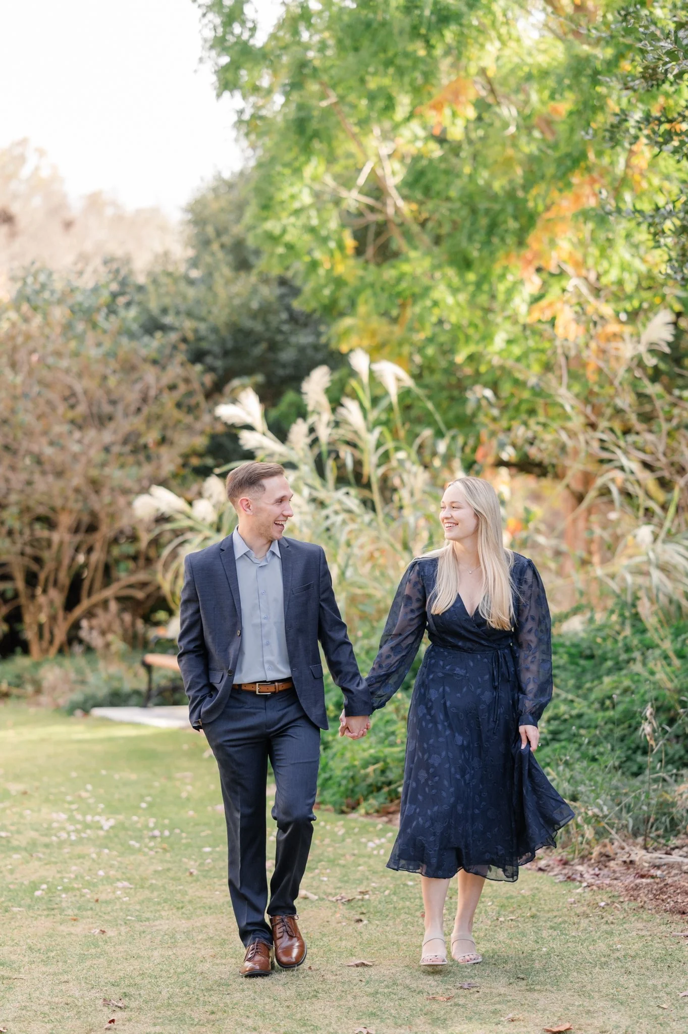 A couple walking hand in hand in a garden, dressed in formal attire; the man is in a dark suit, and the woman is in a dark dress. They are smiling and surrounded by greenery.