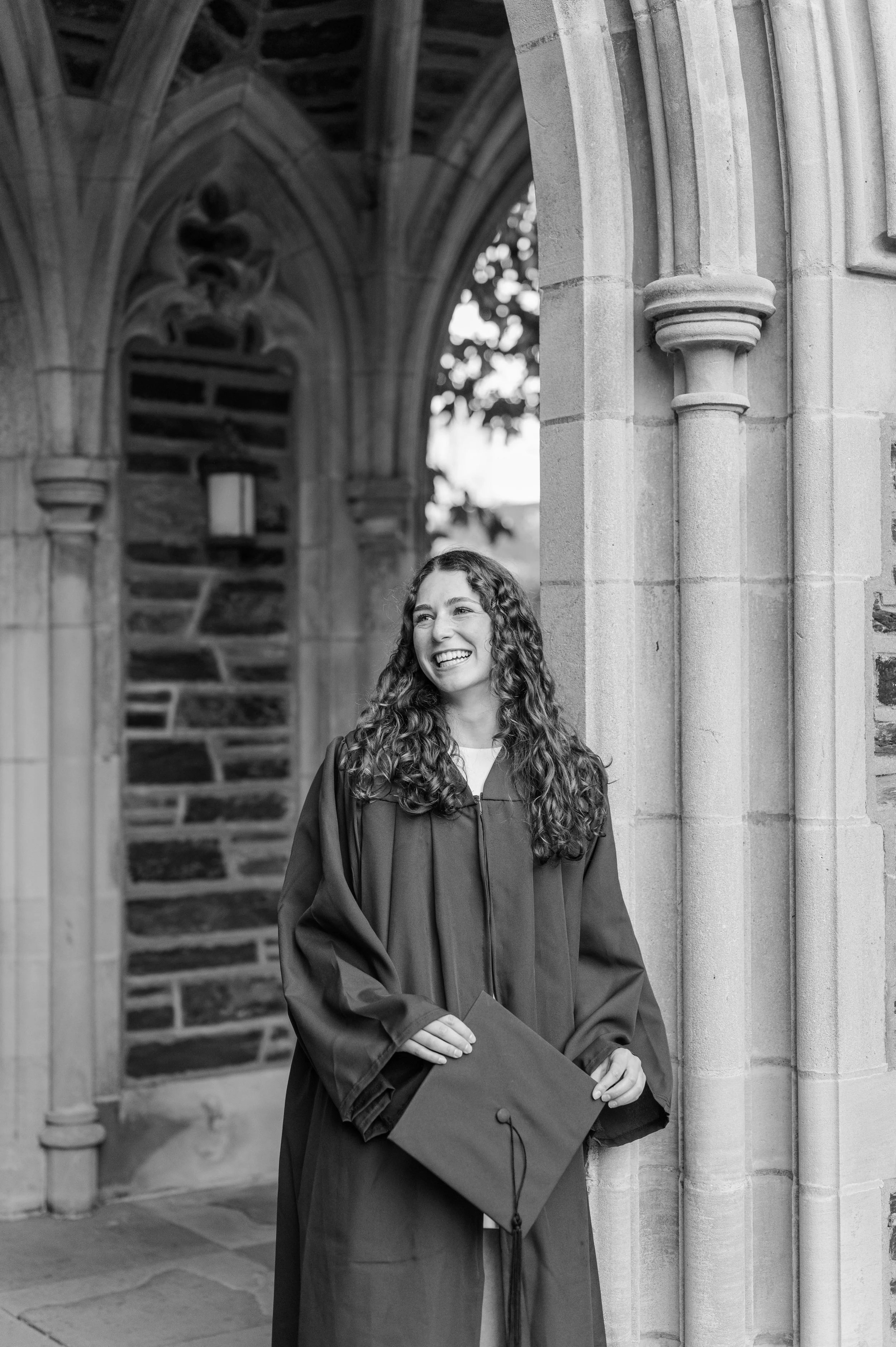 A young woman with long, curly hair smiling in her graduation gown, holding a diploma, standing near the stone architecture of a historic building, possibly a university, during the day.