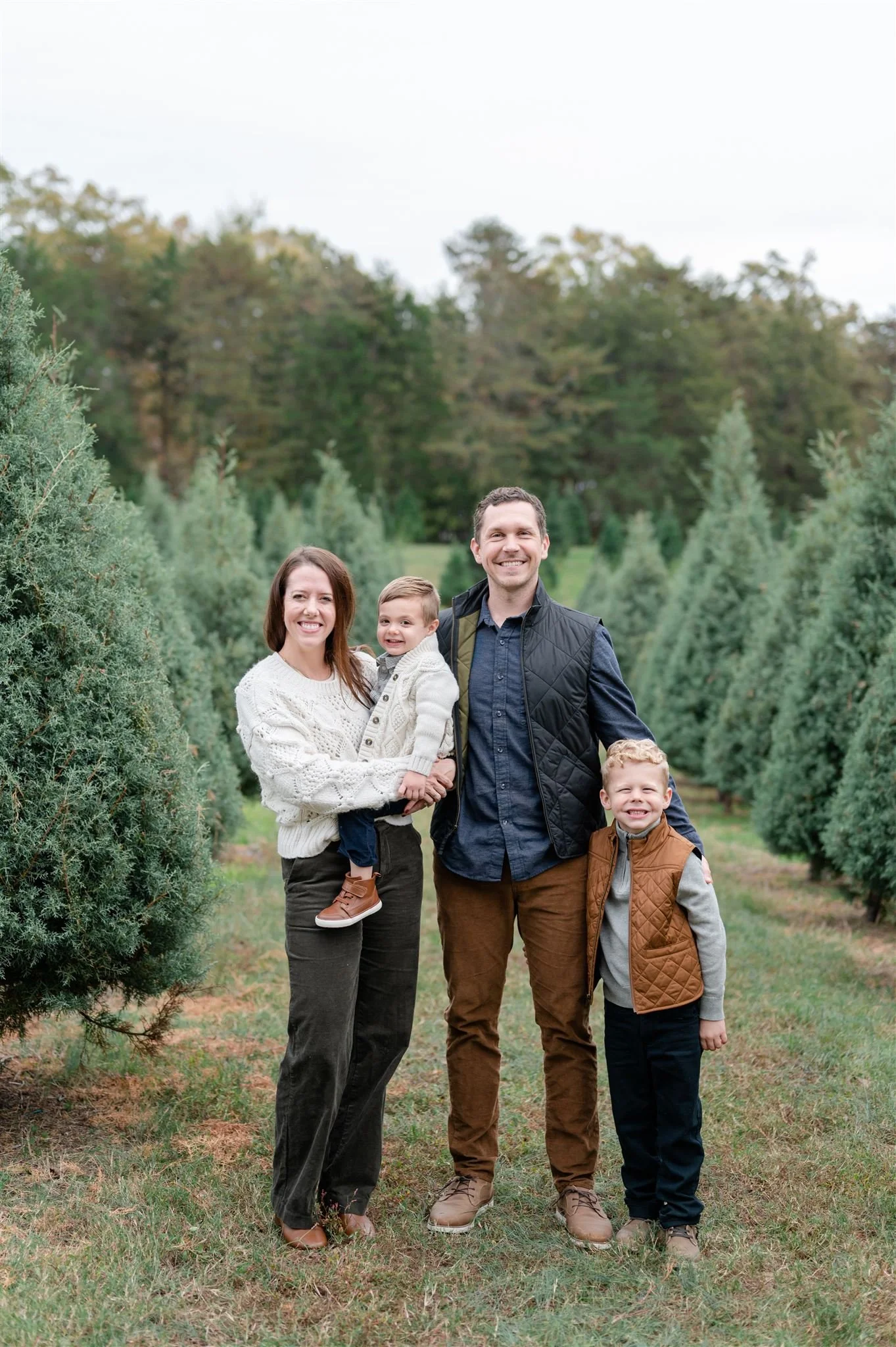 A smiling family of four standing outdoors among Christmas trees in a field, with a backdrop of trees and an overcast sky.