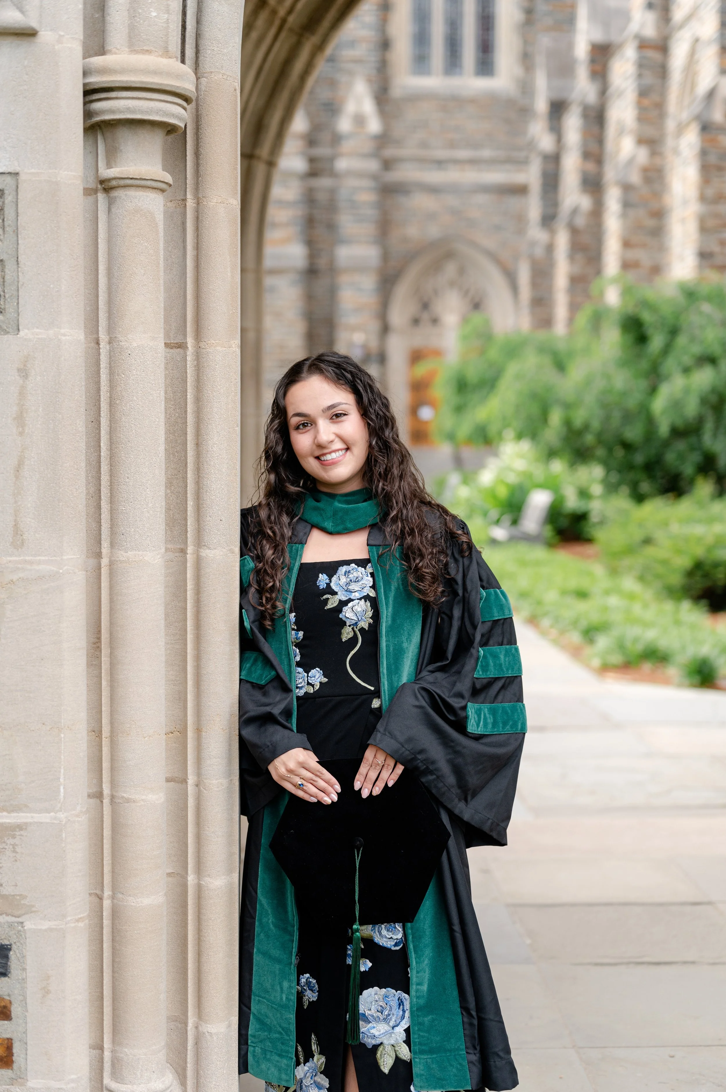 Young woman dressed in a graduation gown with green accents poses outdoors near a stone building with gothic architectural details and greenery in the background.