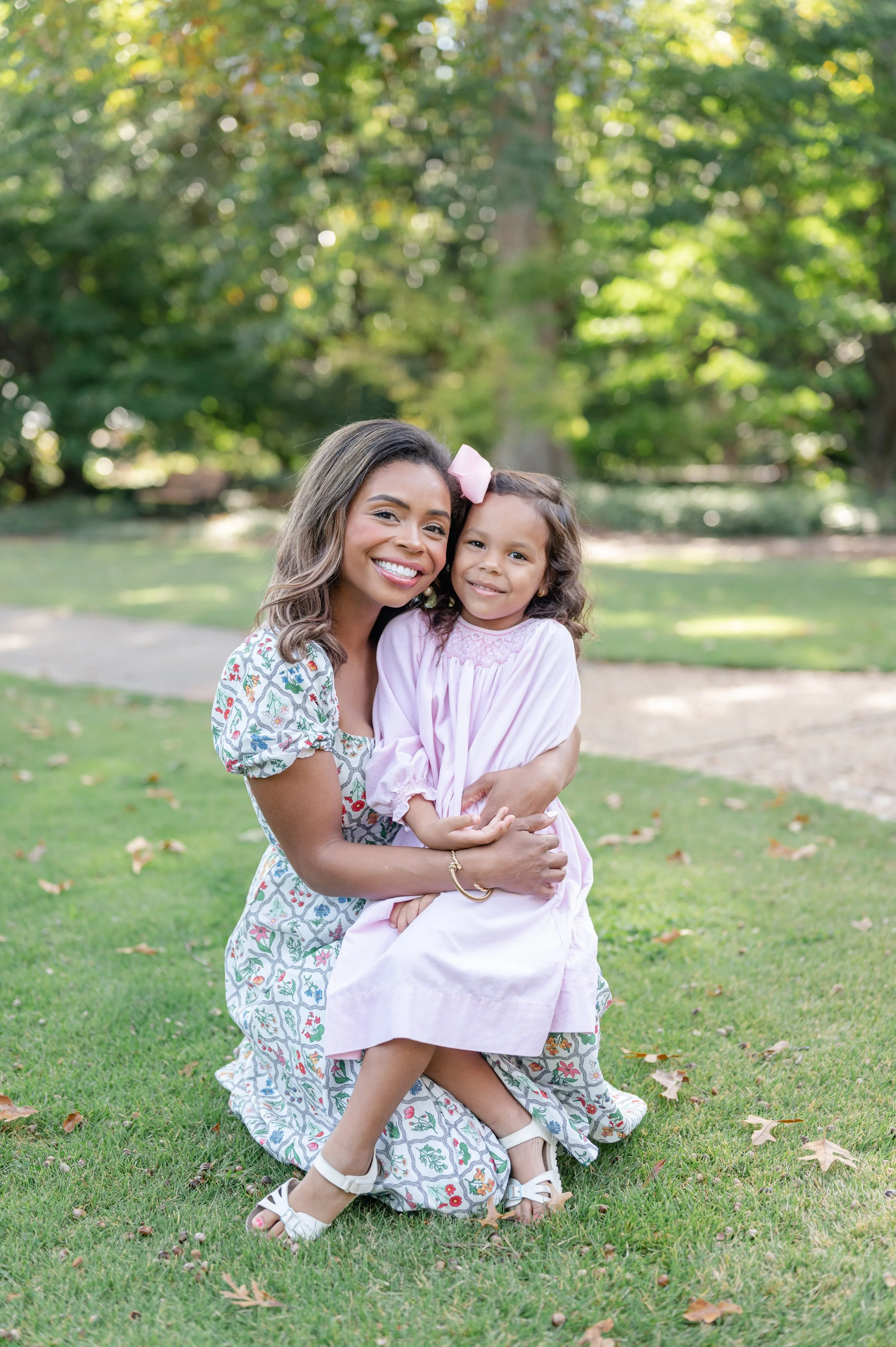 A black woman and a young girl are hugging outdoors in a park with green grass and trees in the background. The woman is wearing a floral dress, while the girl is dressed in a pink dress. Both are smiling at the camera.