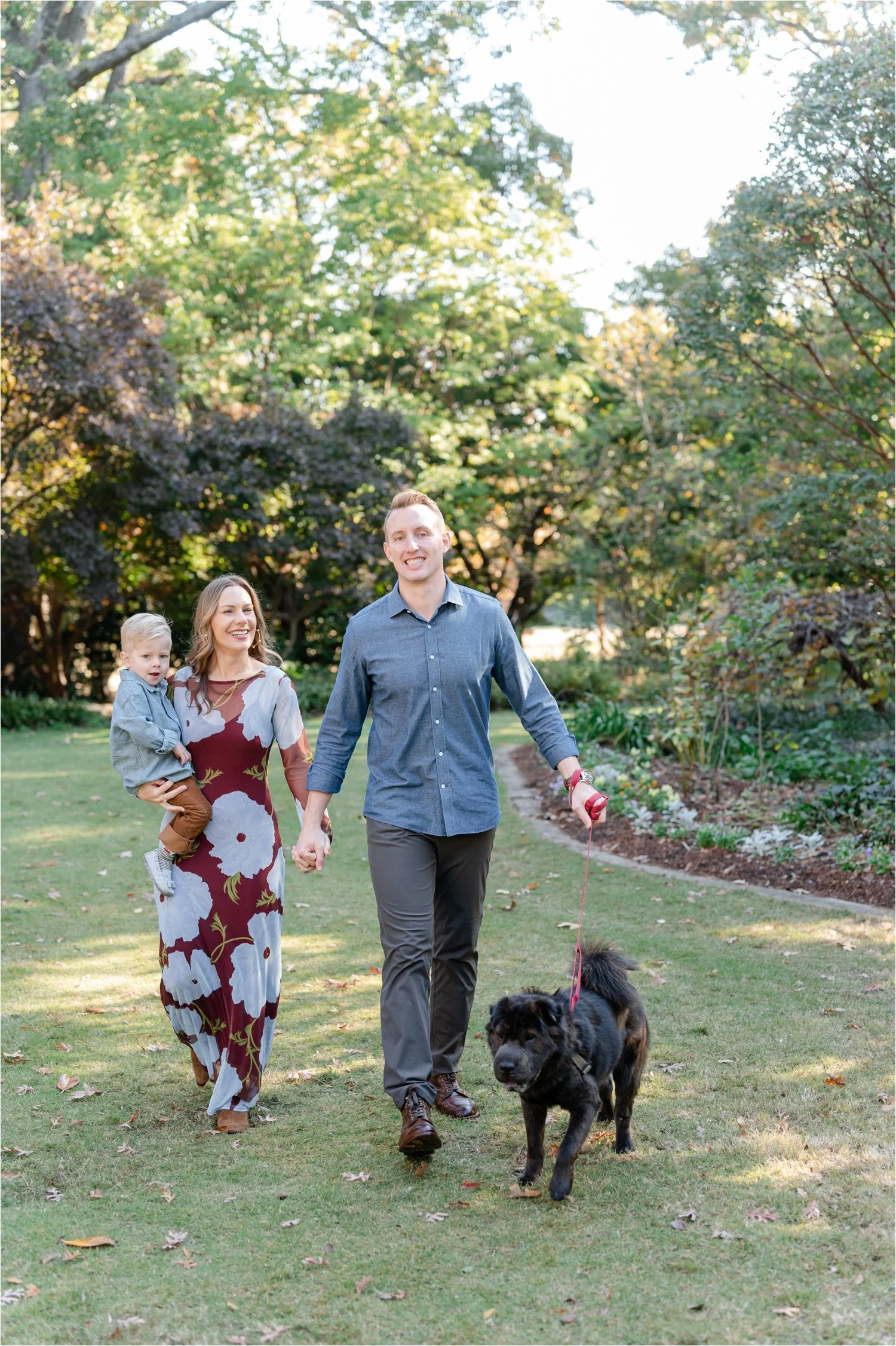 A family of three walking in a park with green trees, a woman holding a young boy, the man holding a dog on a leash, during daytime.