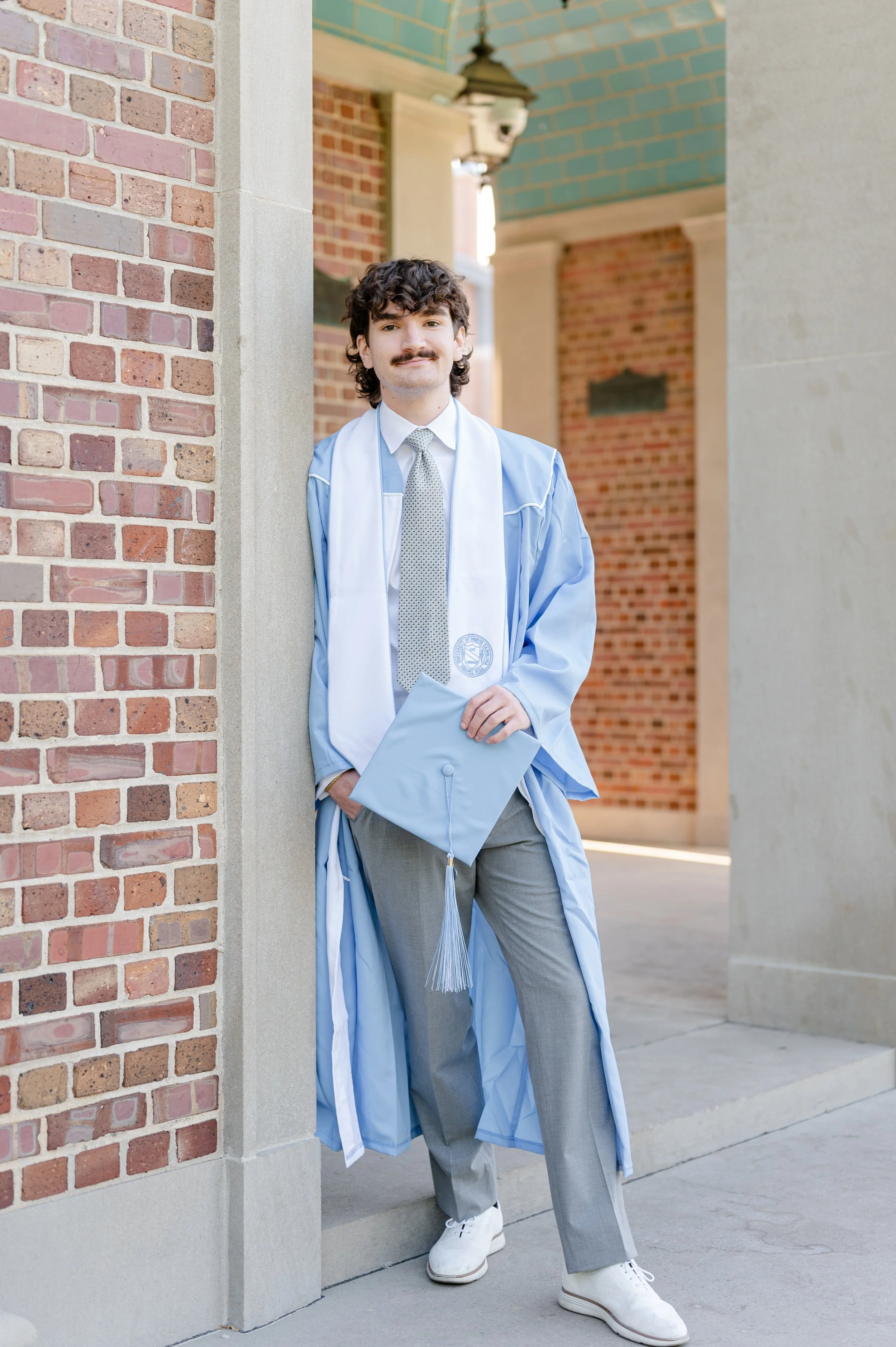A young man in a graduation gown with a cap in hand, leaning against a brick wall outdoors.