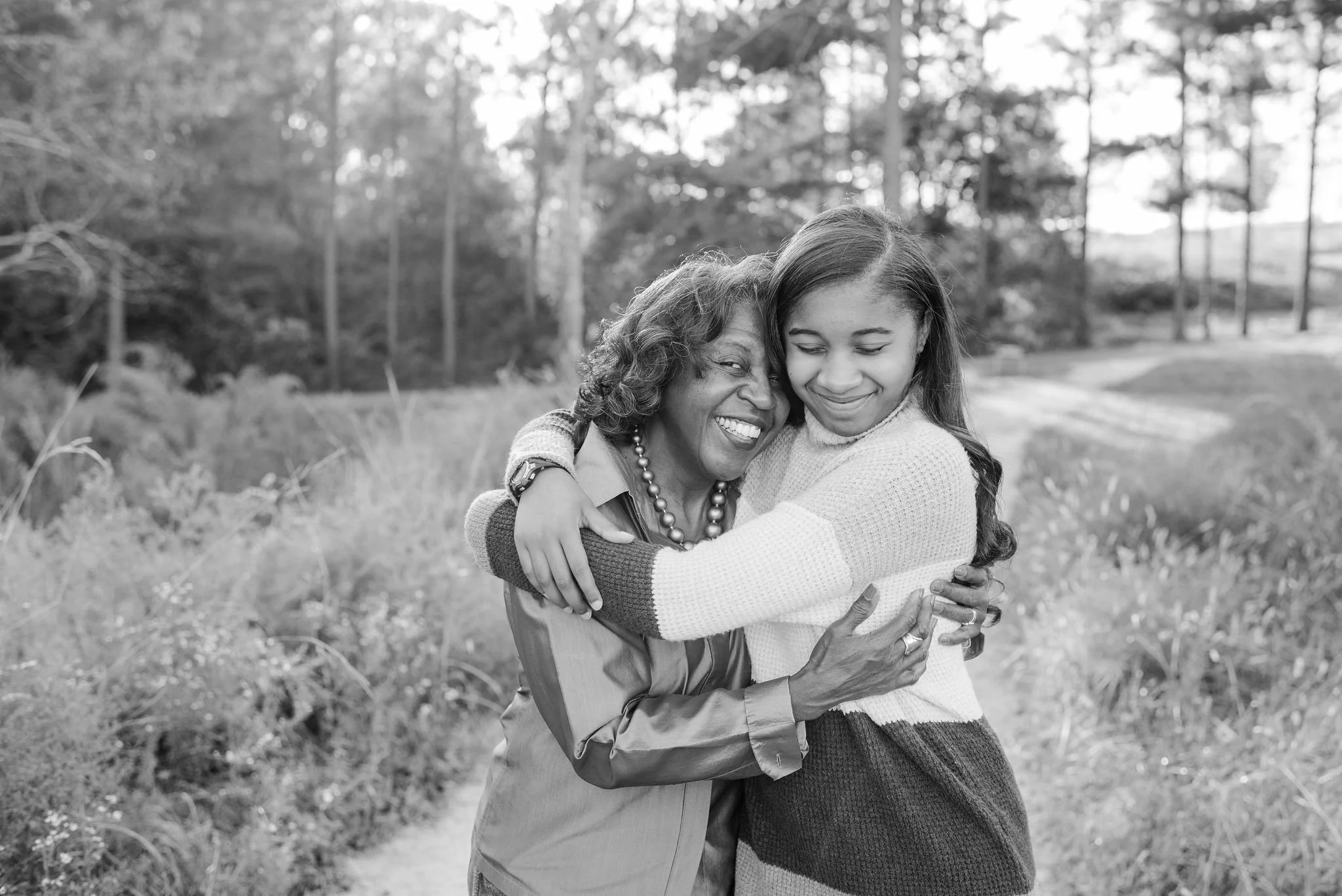 A black woman and her grandaughter hugging outdoors in a park with trees in the background.