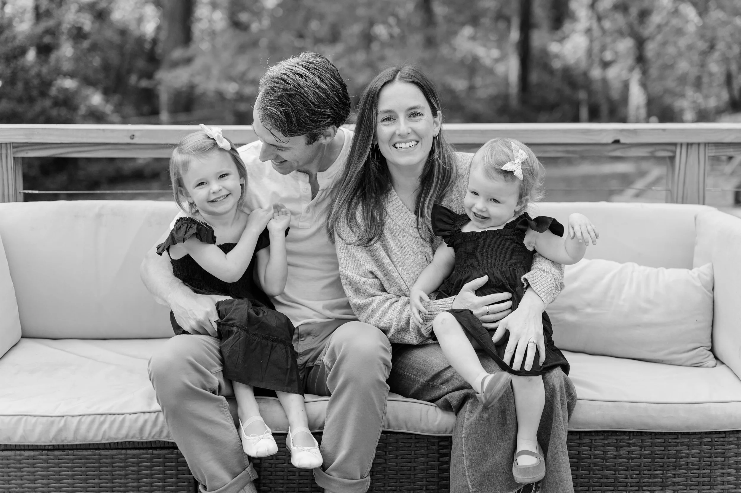 Black and white photo of a family sitting on an outdoor couch. Two young girls and two adults, a man and a woman, are smiling and posing together.