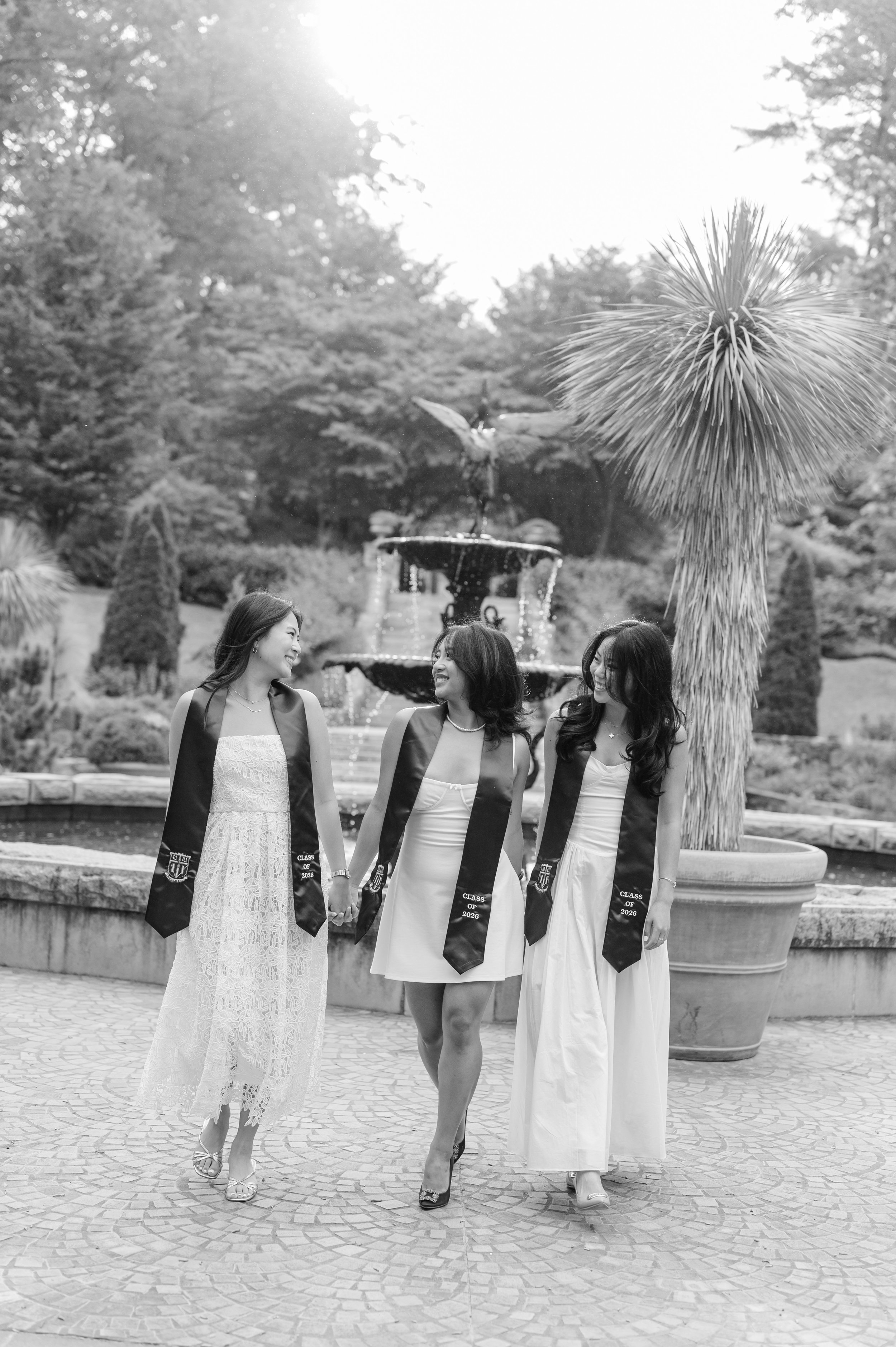 Four women dressed in white dresses and graduation cords, holding hands, walking outdoors near a fountain and large potted plants, celebrating graduation wearing sashes that read "Class of 2026".