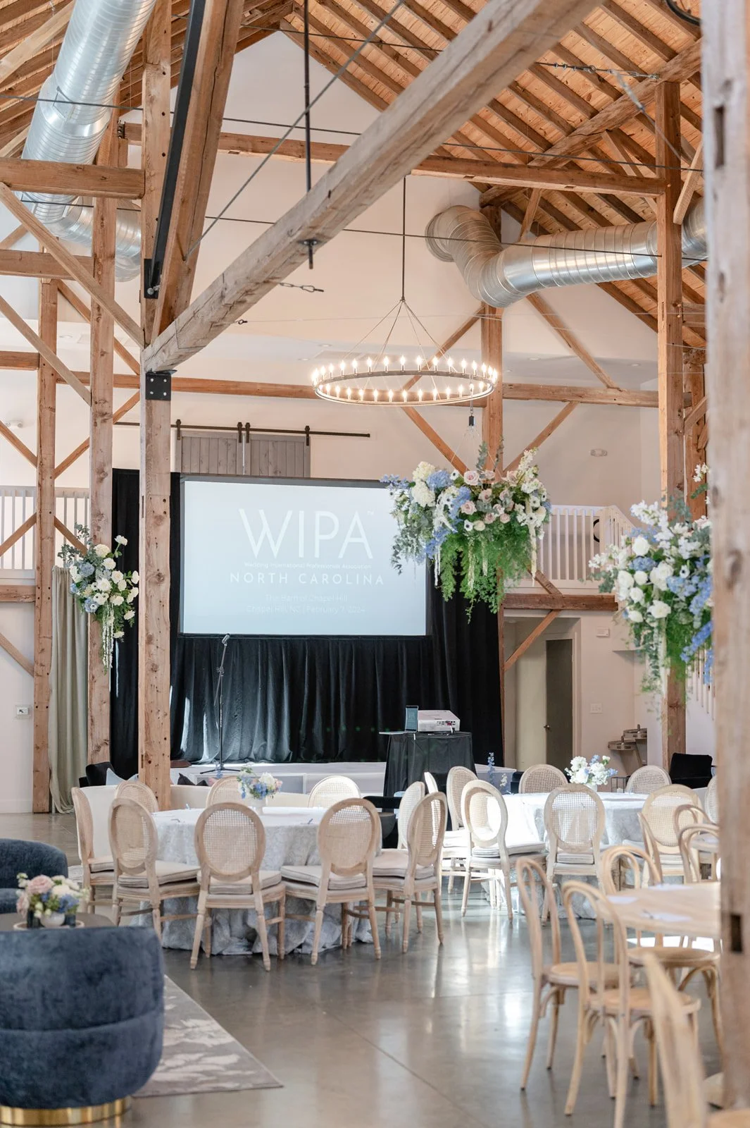 Indoor event space decorated with floral arrangements, featuring a large screen with 'WIPA North Carolina' displayed, surrounded by rows of chairs and tables for a gathering or conference.