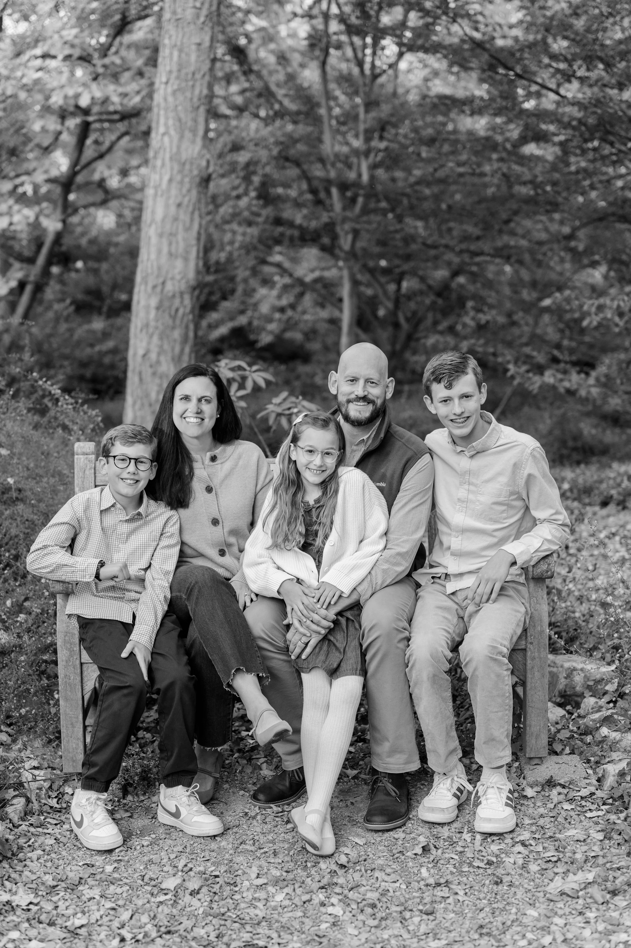 A family of six sitting outdoors on a wooden bench, smiling at the camera, with trees in the background. The family includes two adults and four children, all casually dressed.