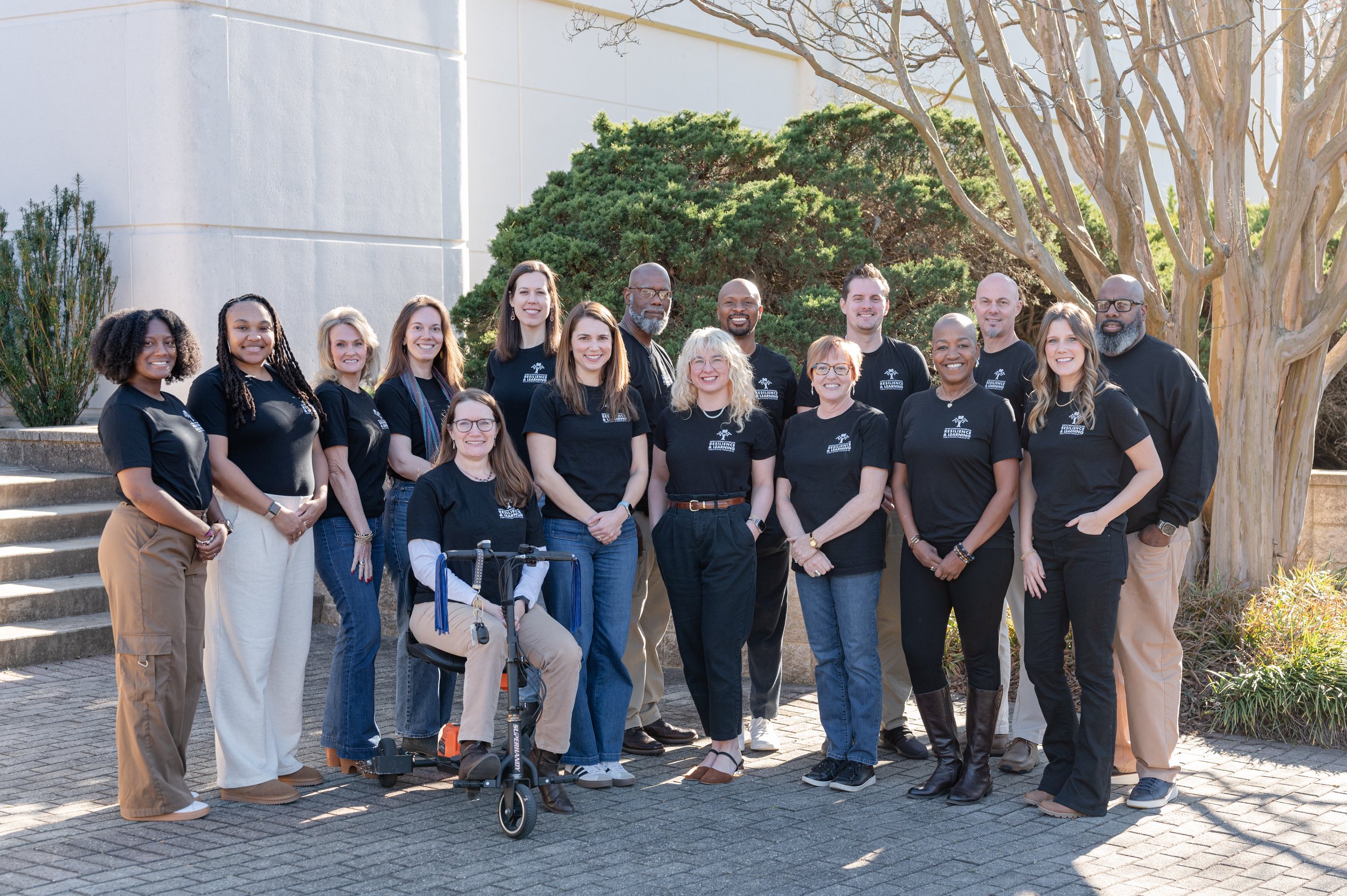 Group of diverse people standing outdoors in front of trees and a building, wearing black T-shirts with a logo, smiling for a photo.