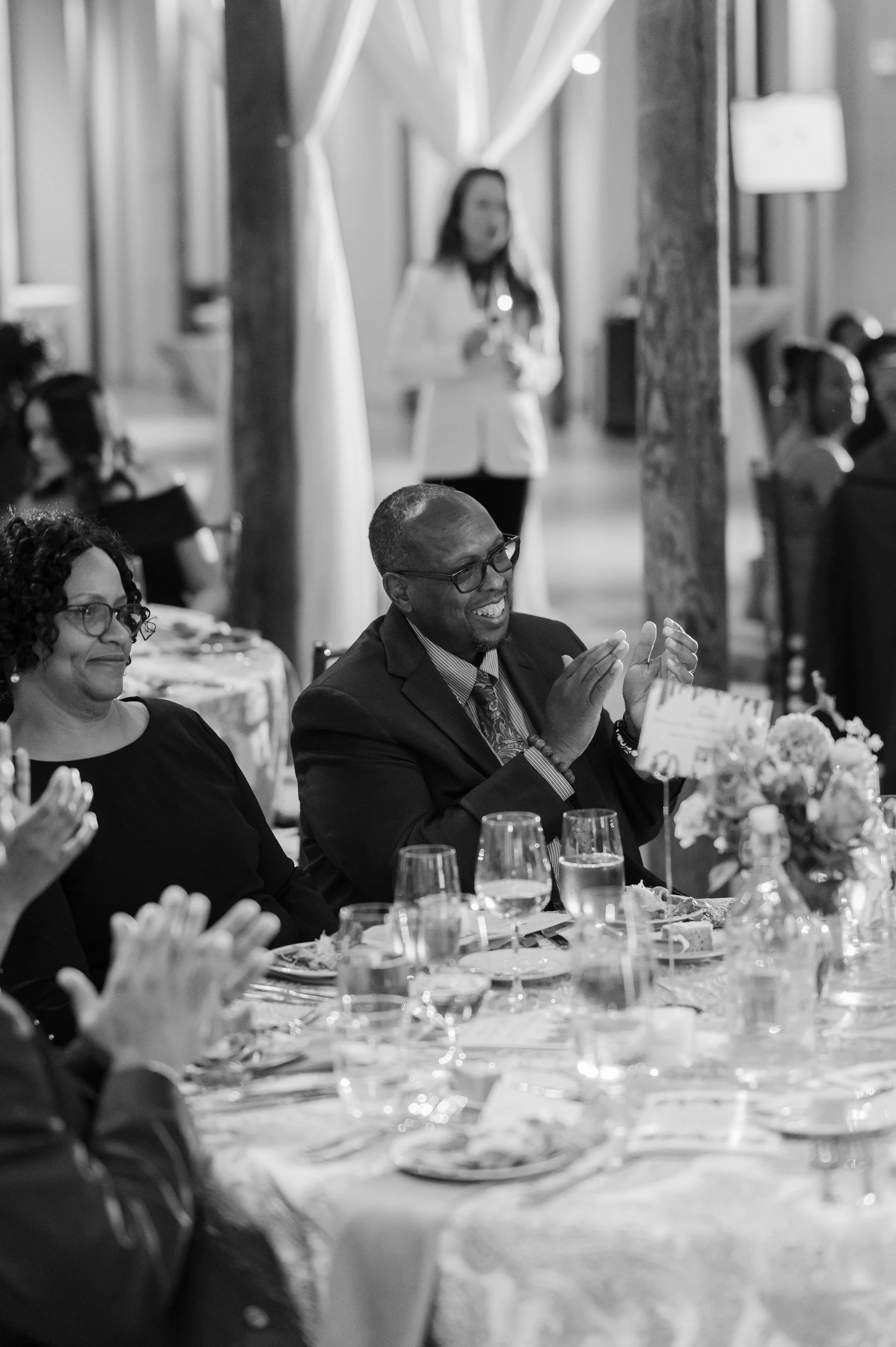 People seated at a banquet table clapping and smiling during a formal event, with a woman standing in the background.