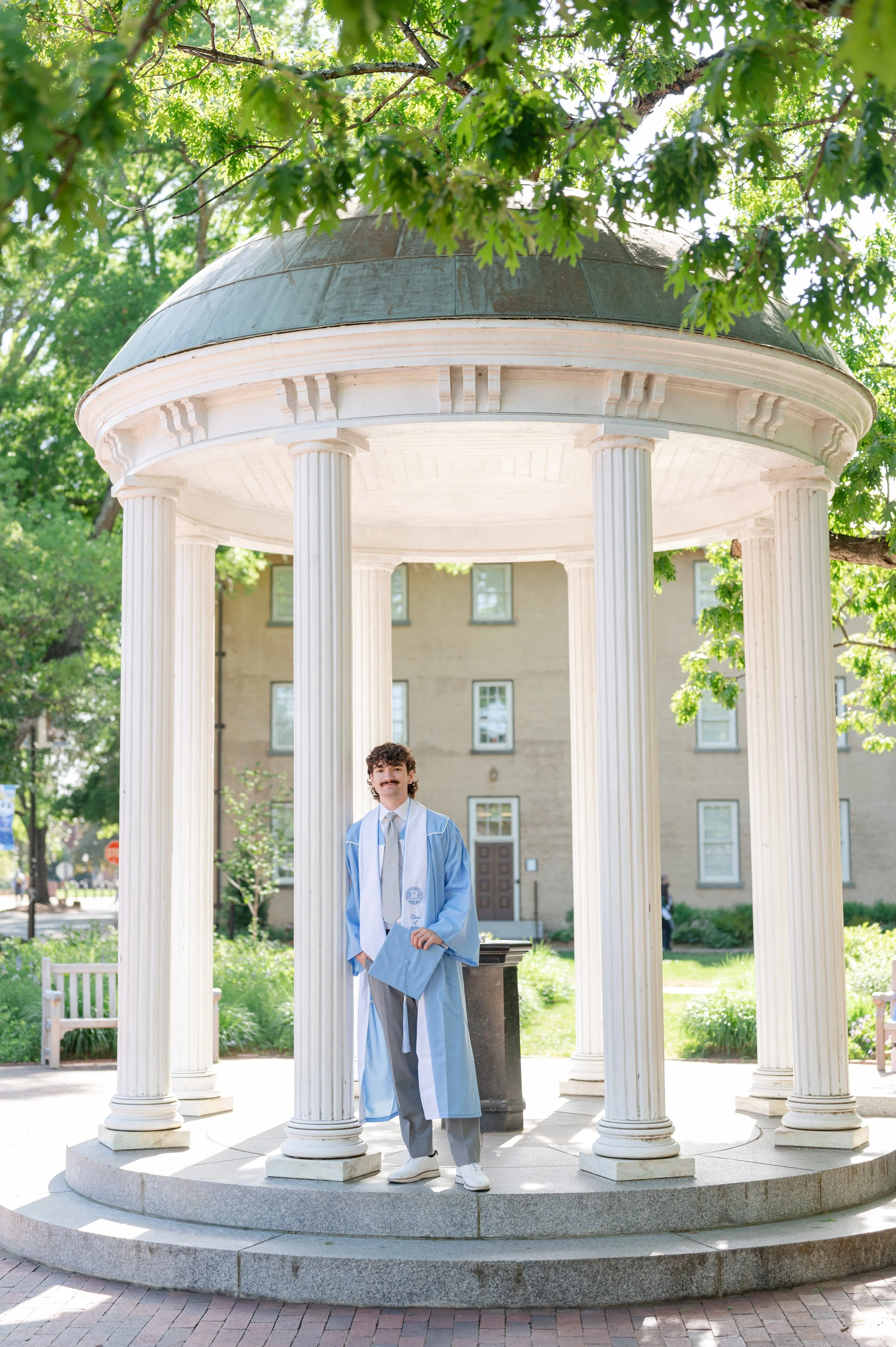 A young man in a light blue graduation gown and cap standing on the steps of a white, classical pavilion with columns, holding his diploma, in a park with greenery and trees.