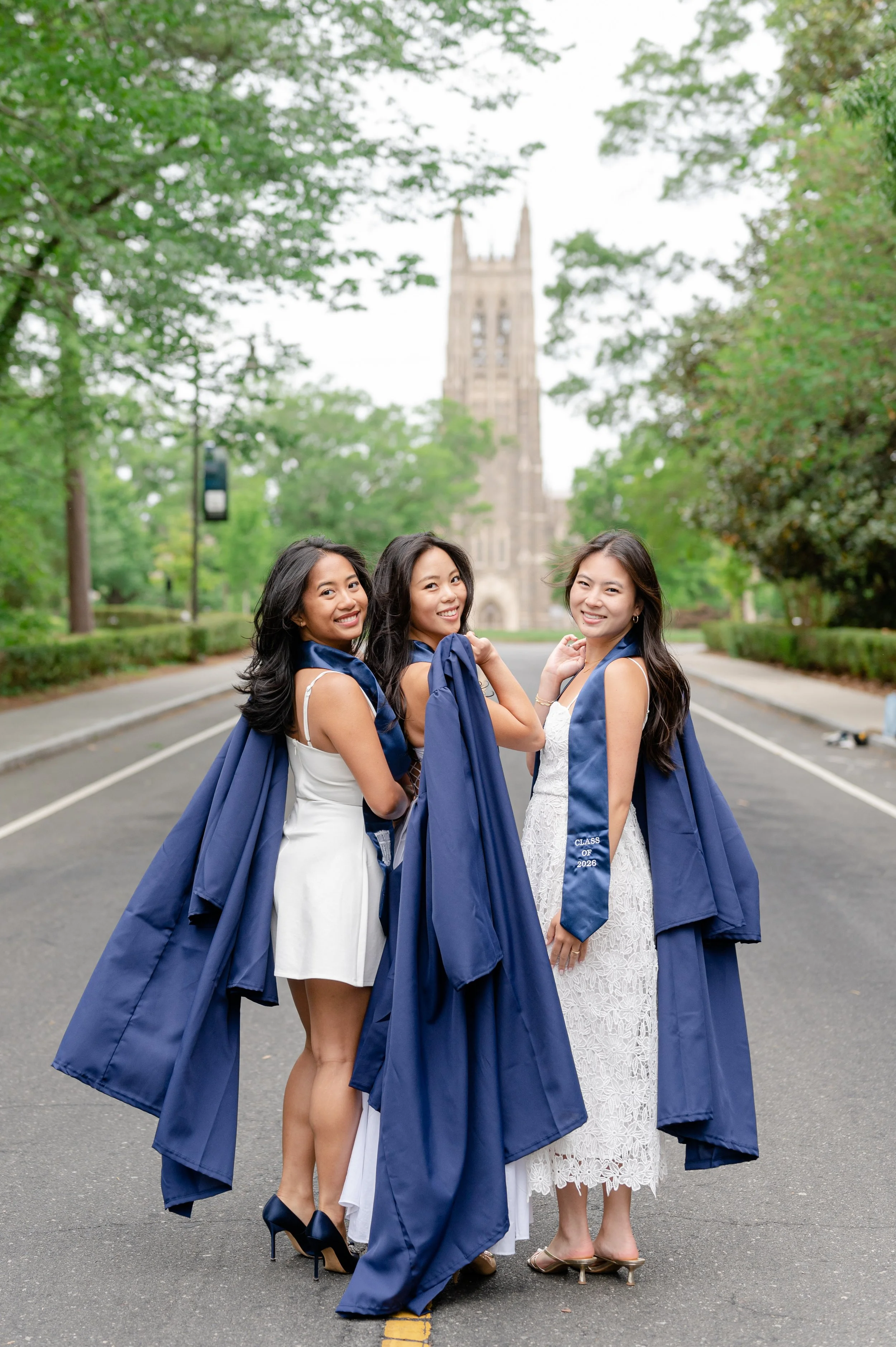Three women in graduation gowns and dresses standing on a street with a university building with a bell tower in the background, smiling at the camera.