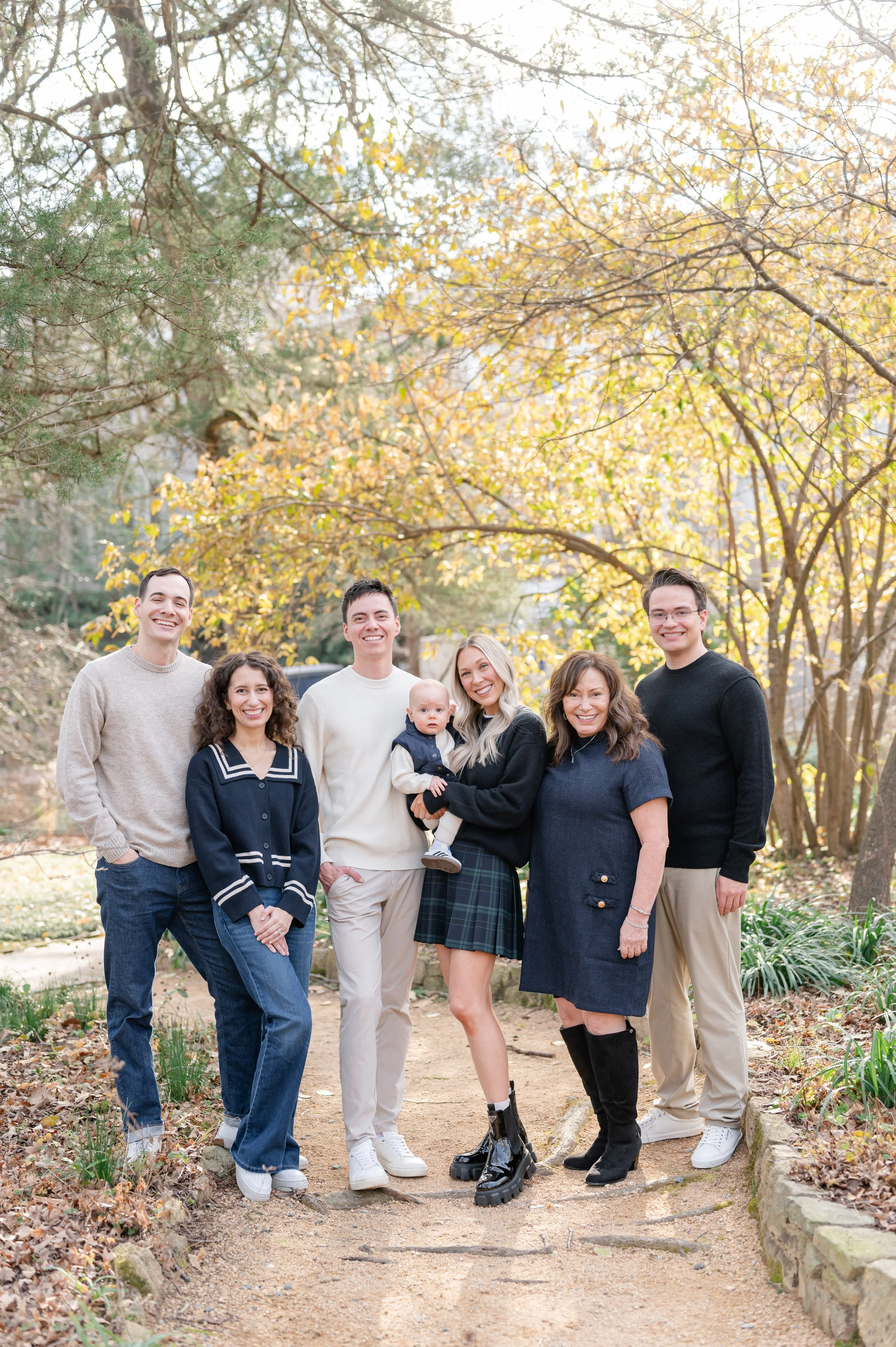 Group of seven people standing outdoors in fall, smiling, in a park with yellow and orange trees.