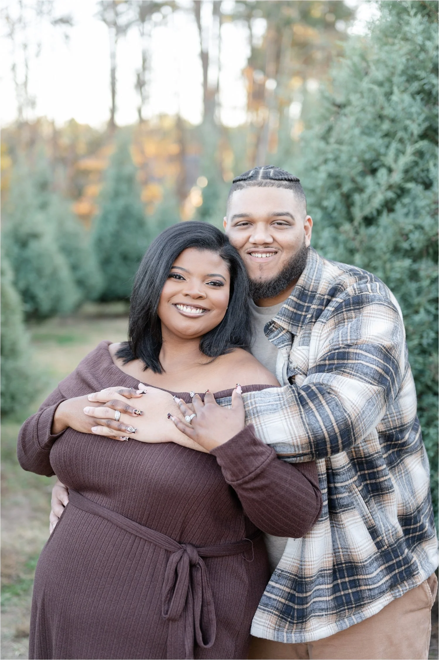 A smiling black couple, with the woman wearing a brown dress and the man in a plaid shirt, embracing outdoors with trees in the background.