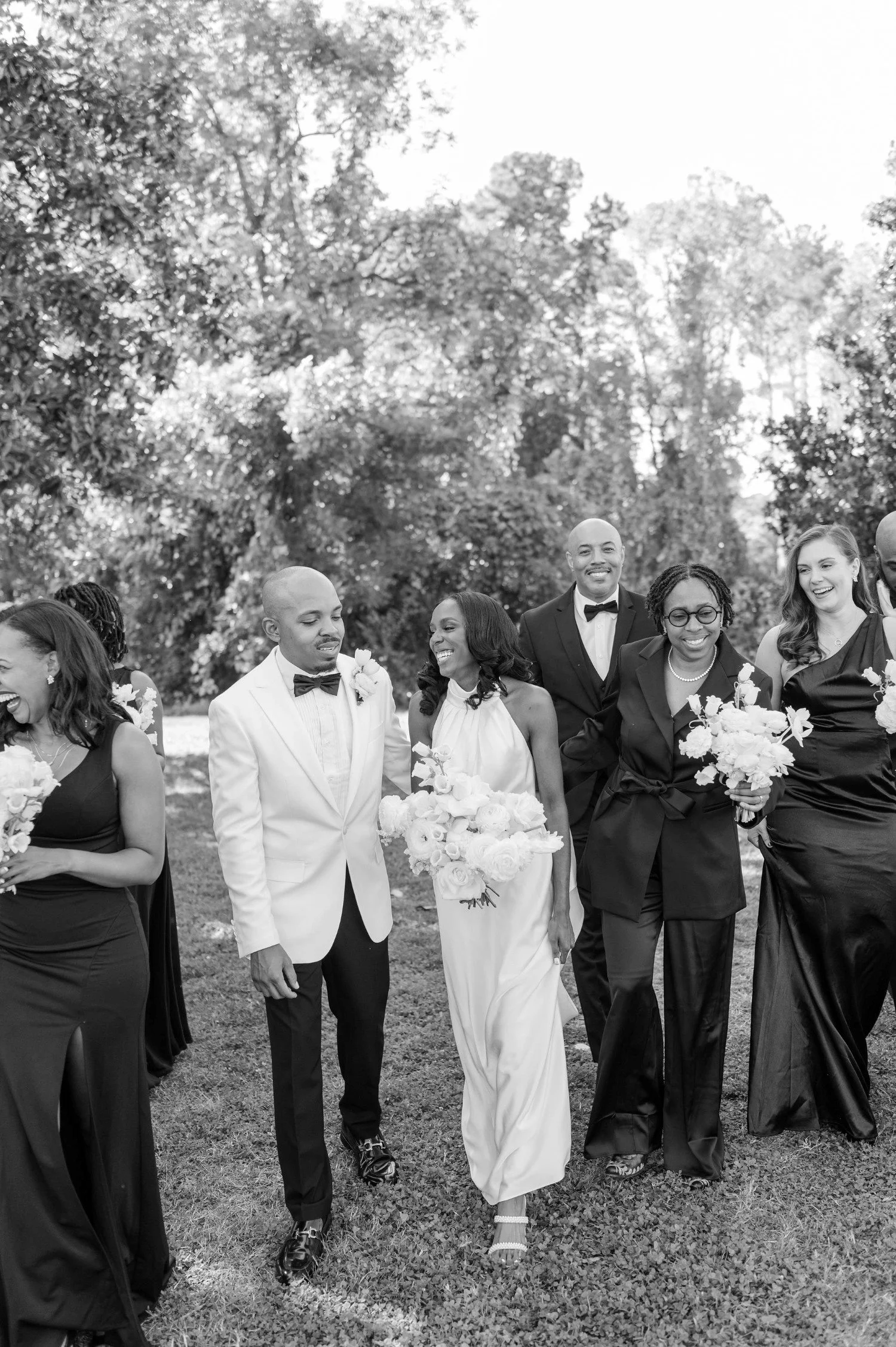 A black and white photo of a wedding party. The group includes a bride in a white gown holding a bouquet, a groom in a white tuxedo jacket with a bow tie, and several bridesmaids and groomsmen dressed in black attire, walking and smiling outdoors with trees in the background.