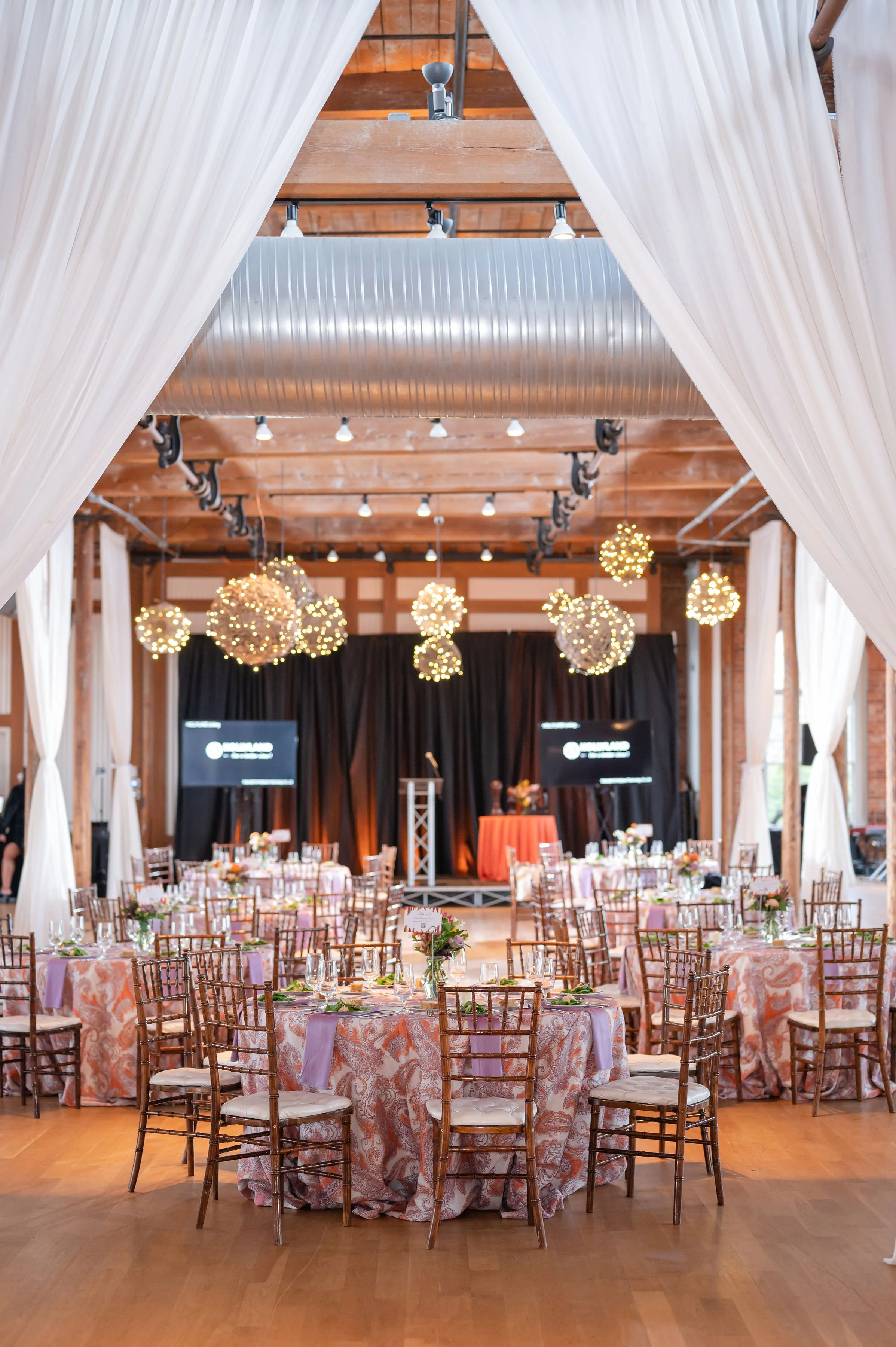 A banquet hall decorated for an event with round tables covered in pink and orange patterned tablecloths, floral centerpieces, and wooden chairs. The room features draped white curtains, string lights, and a stage area with black curtains and monitors.