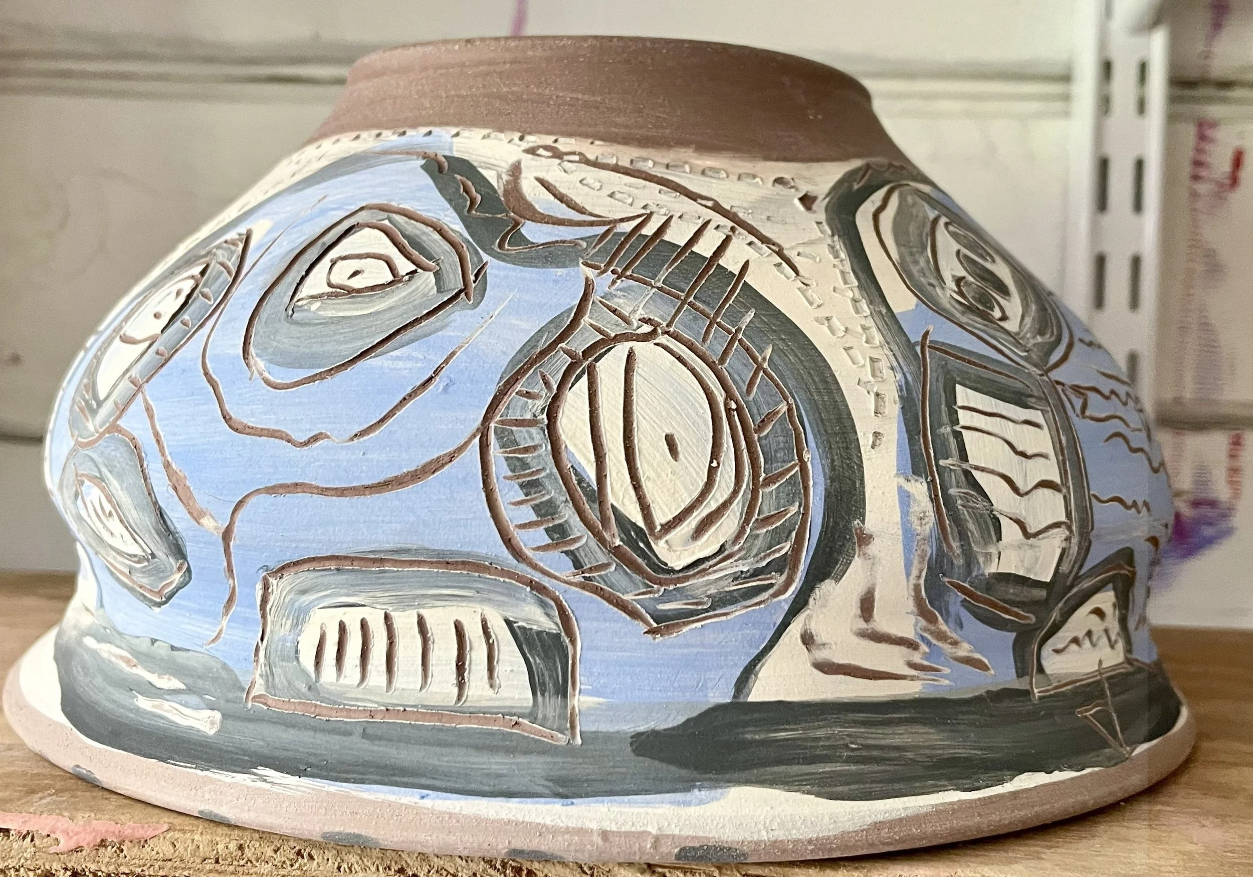 Painted ceramic object with abstract geometric and face-like designs in black, gray, white, and blue on a wooden surface.