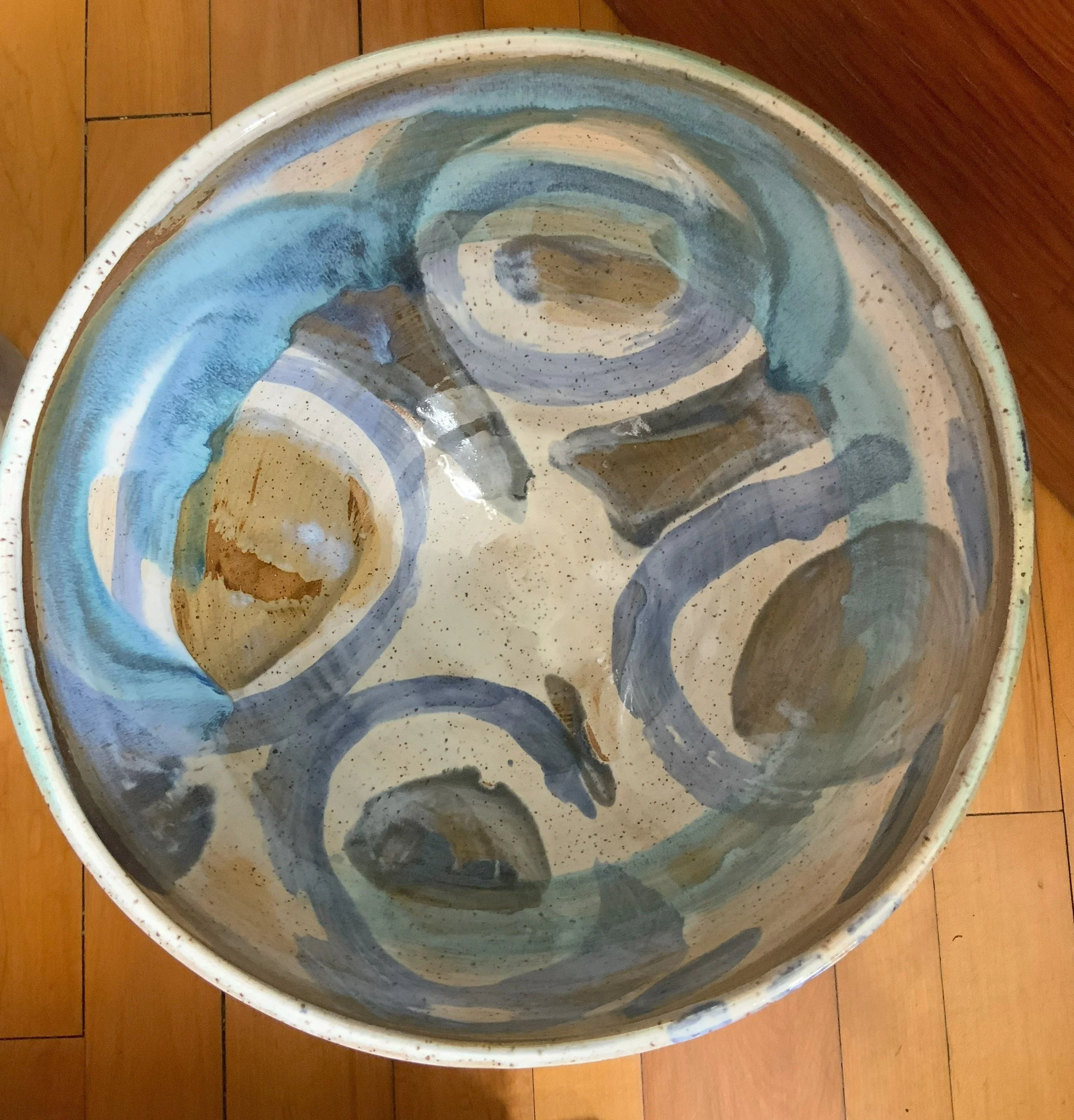 A large ceramic bowl with abstract blue, brown, and beige swirl patterns along the inside rim, placed on a wooden floor.