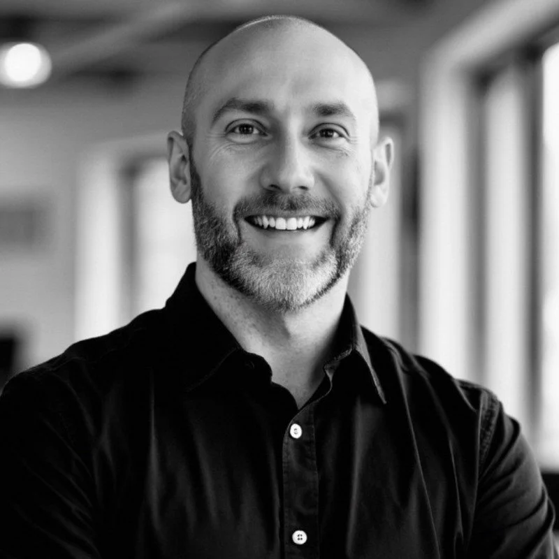Black and white portrait of a bald man with a beard, smiling, wearing a dark shirt, in a well-lit indoor setting.
