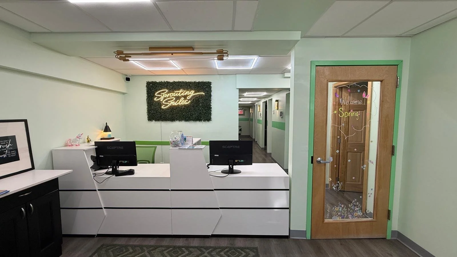 Reception area with partial view of the coffee counter with a picture frame on it, a white desk, two computer monitors, an office chair, green plant wall with neon sign that says "sprouting smiles", and a hallway with glass door and green trim.
