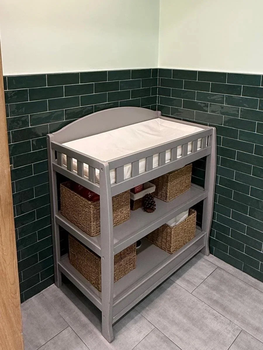 grey changing table with 2 shelves and 5 containersin corner of bathroom in front of green subway tiles 