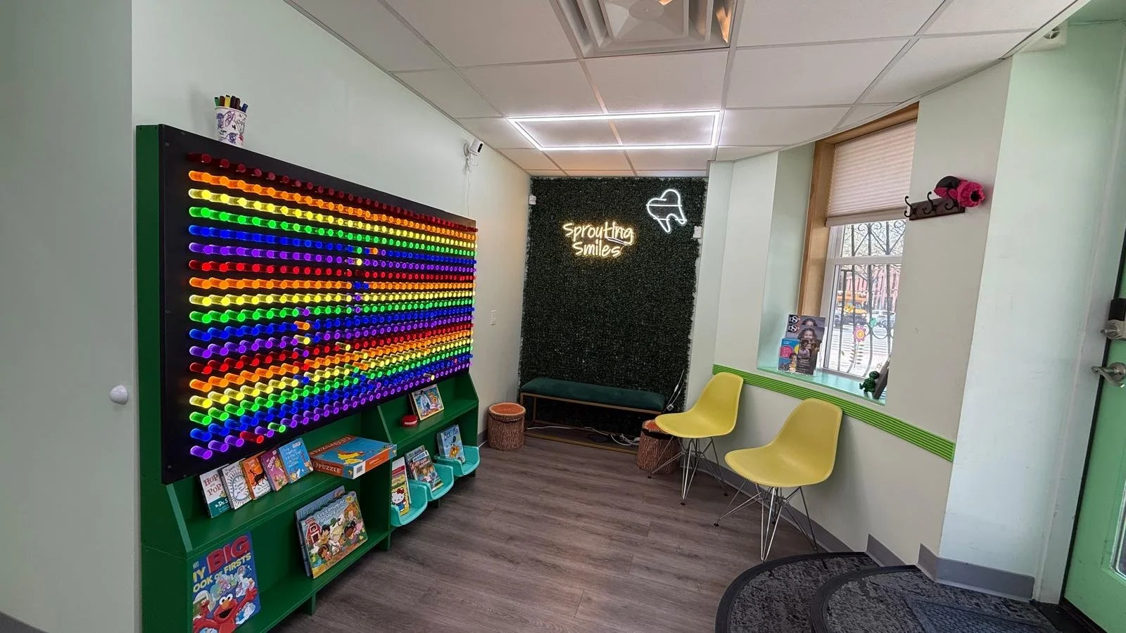 Colorful light board with books below, two empty yellow chairs, small green bench , neon sign of a tooth and one that says "Sprouting Smiles" on a green wall with artificial plants.