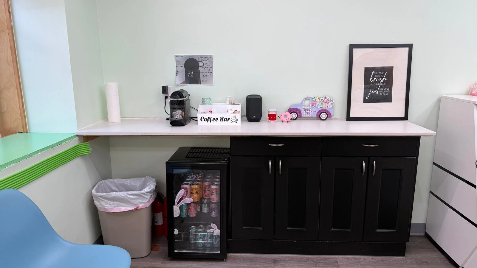 coffee station: white counter along wall, roll of paper towel, coffee machine + supplies, speaker, decorations (felt coffee, donut, flat wooden purple flower truck, framed quote (words not visible), black cabinet, small fridge, garbage under counter