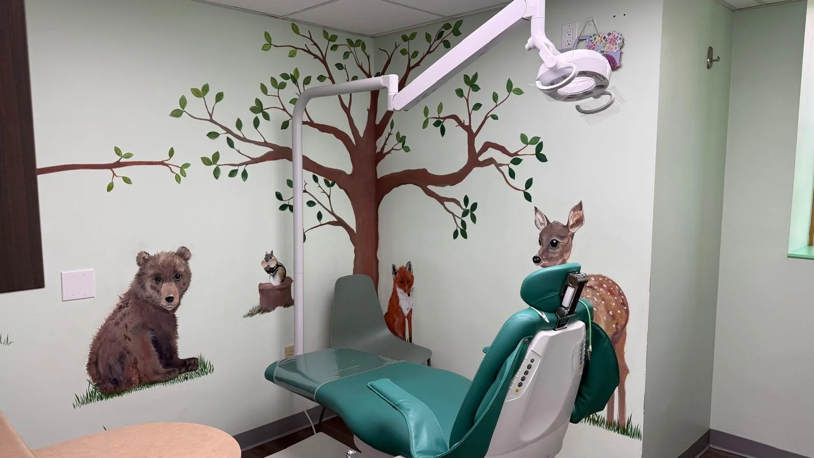 operatory: dental chair, mural on the wall of a tree where the 2 walls meet, branches extend onto each wall with green leaves, a baby deer and fox on right wall, a baby bear and chipmunk on a log on left wall, partial view of desk on left