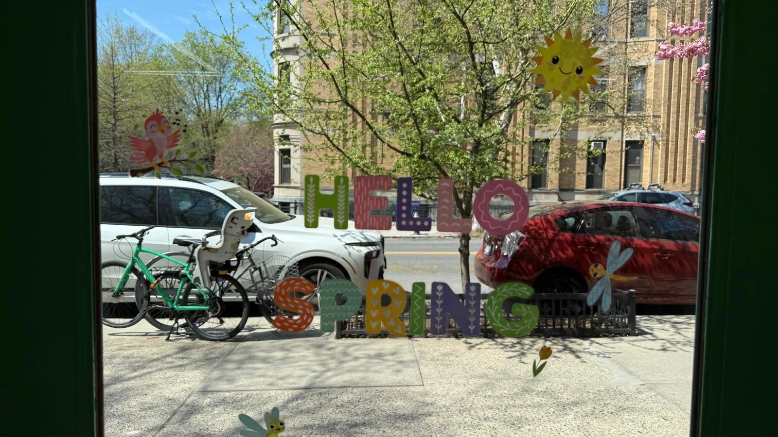 picture of view through glass door showing street with cars, and trees, with "hello spring" glass stickers on door