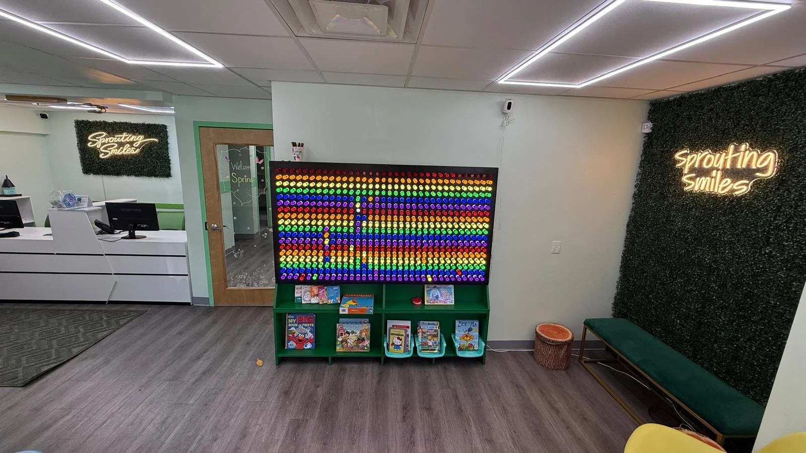 First thing you see when you walk in to the office. View of entire waiting area: light board with books under it, green plant wall with neon sign "sprouting smiles" and green bench to the right, reception to the left with a glass door that leads back