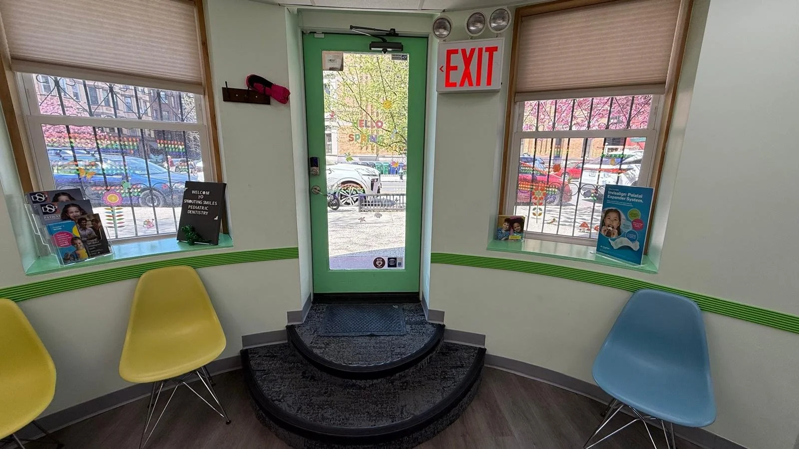 exit: door is center with exit sign to the top right, blue and yellow chairs and windows on either side of glass door with green frame, 2 rounded steps leading to door