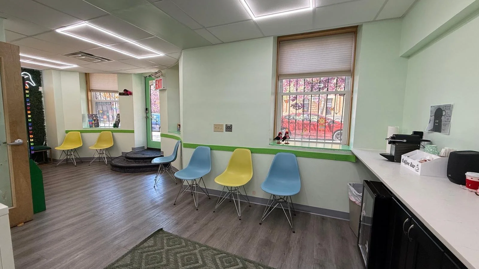 An office waiting area with colorful chairs (yellow and blue) lining the walls on either side of the entrance, 2 windows with open blinds, a coffee station on the right wall, coffee machine and supplies, small fridge underneath counter.
