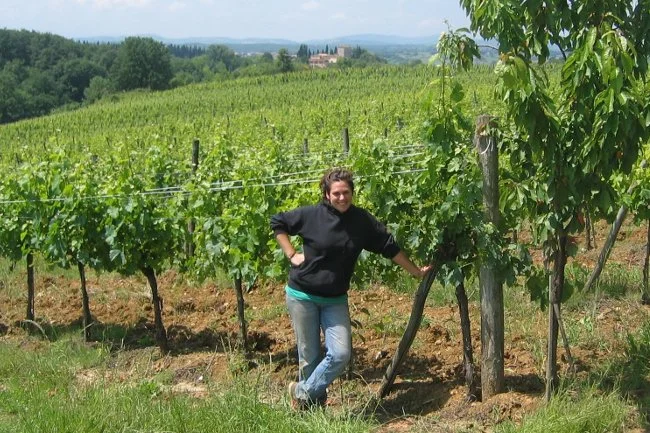 A winemaker standing in a vineyard with green grapevines and a tree, with rolling hills and a building in the background.