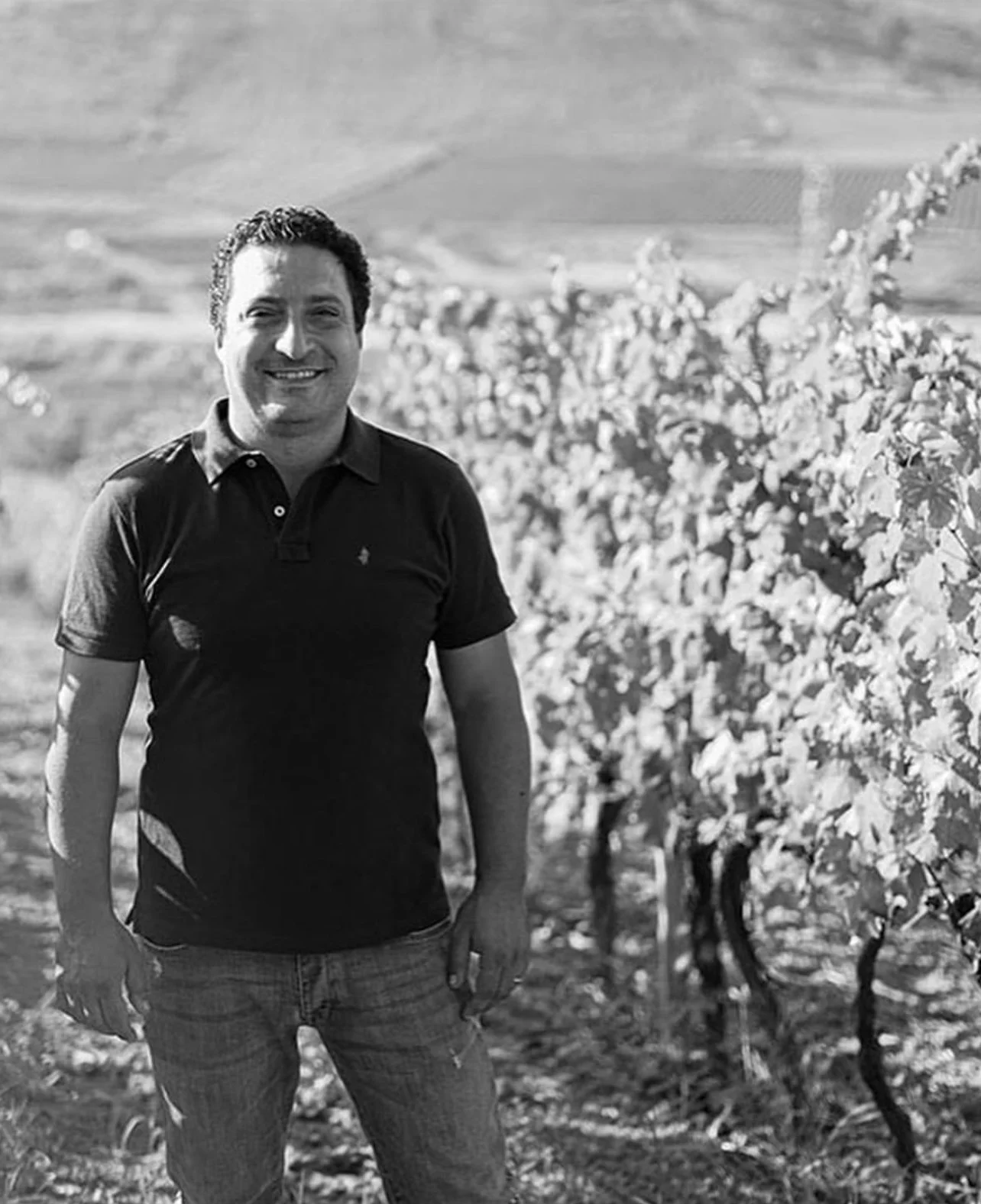 A winemaker smiling and standing in a vineyard with rows of grapevines in the background.