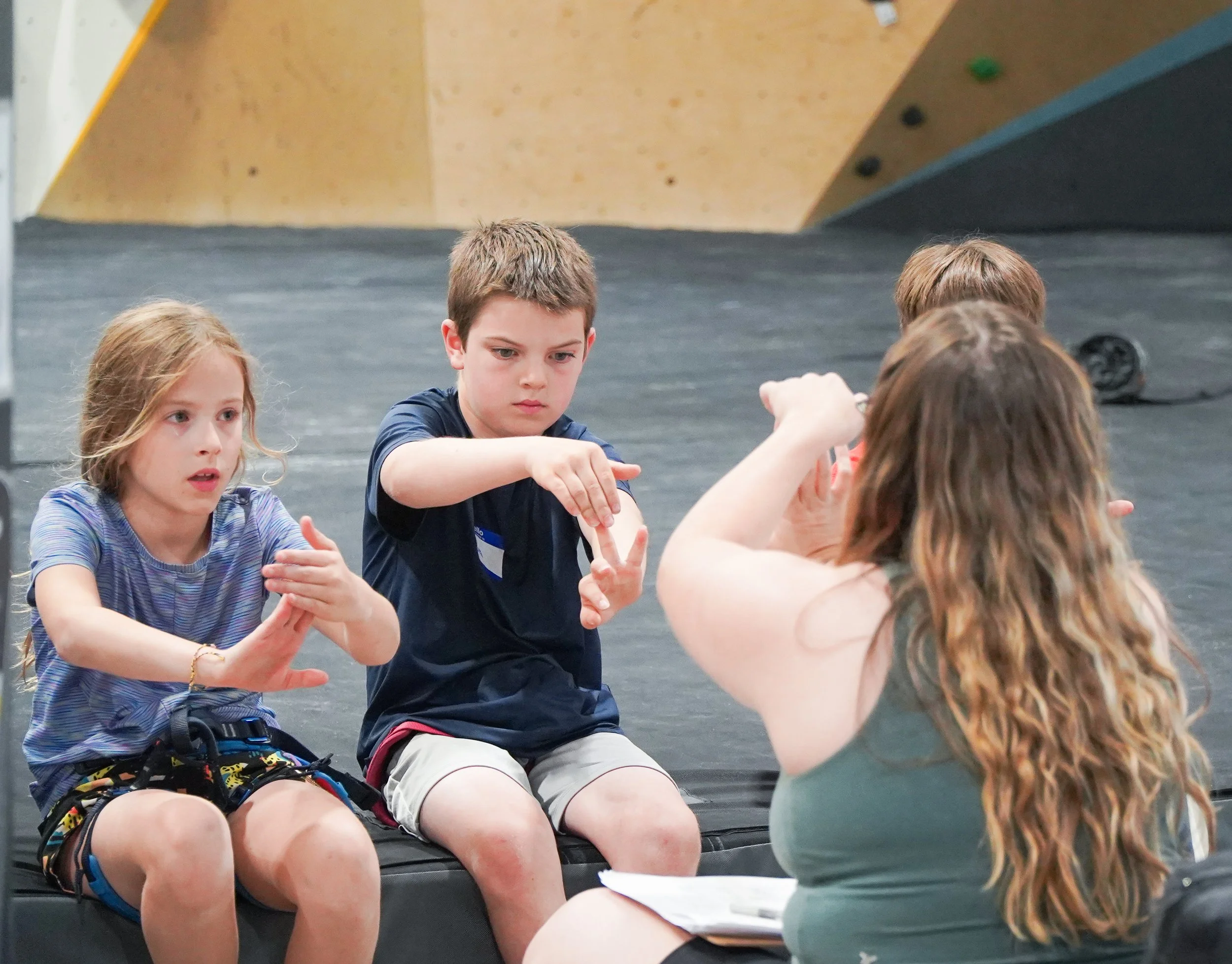 Three children sitting on a black mat listening to a woman with long brown hair who is talking and gesturing with her hands, in what appears to be an indoor climbing gym.