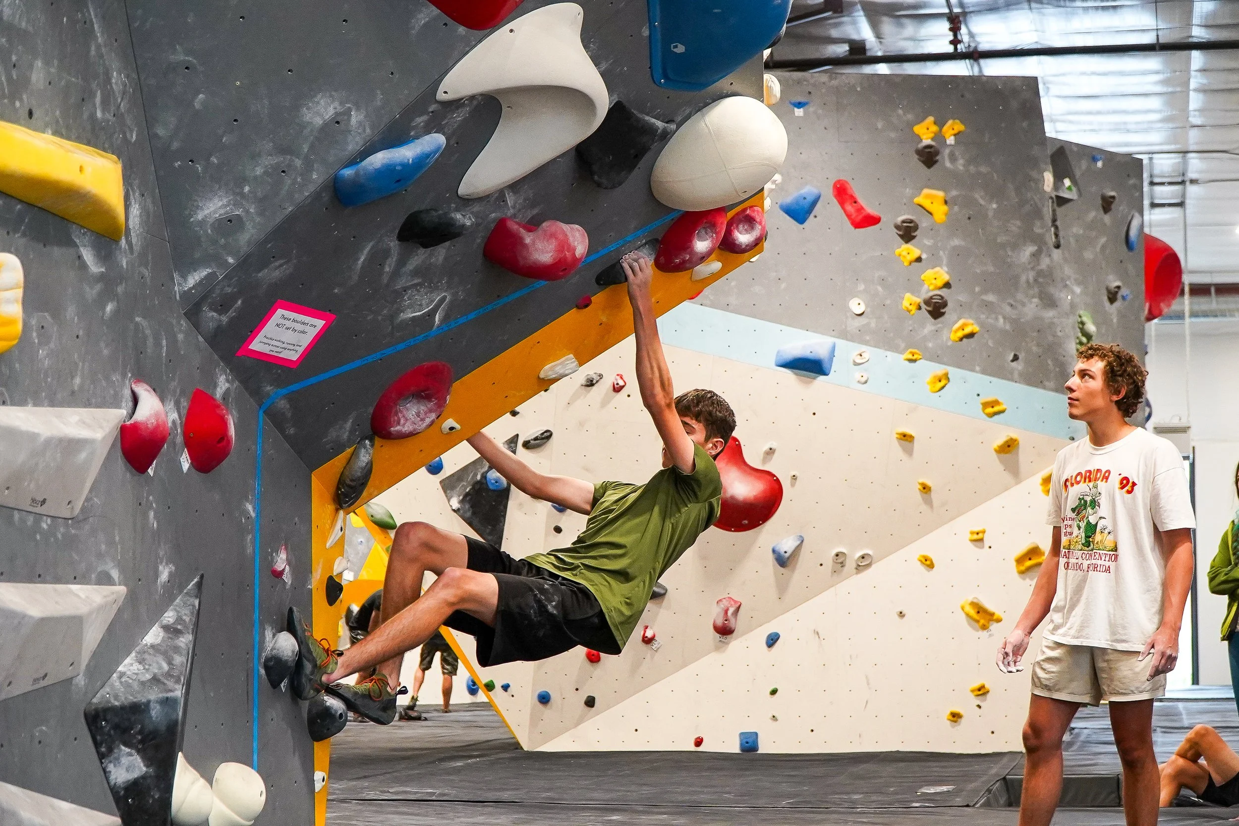 A youth team athlete climbing an overhung boulder. There is a fellow youth team athlete watching nearby as the climber goes for their next move.