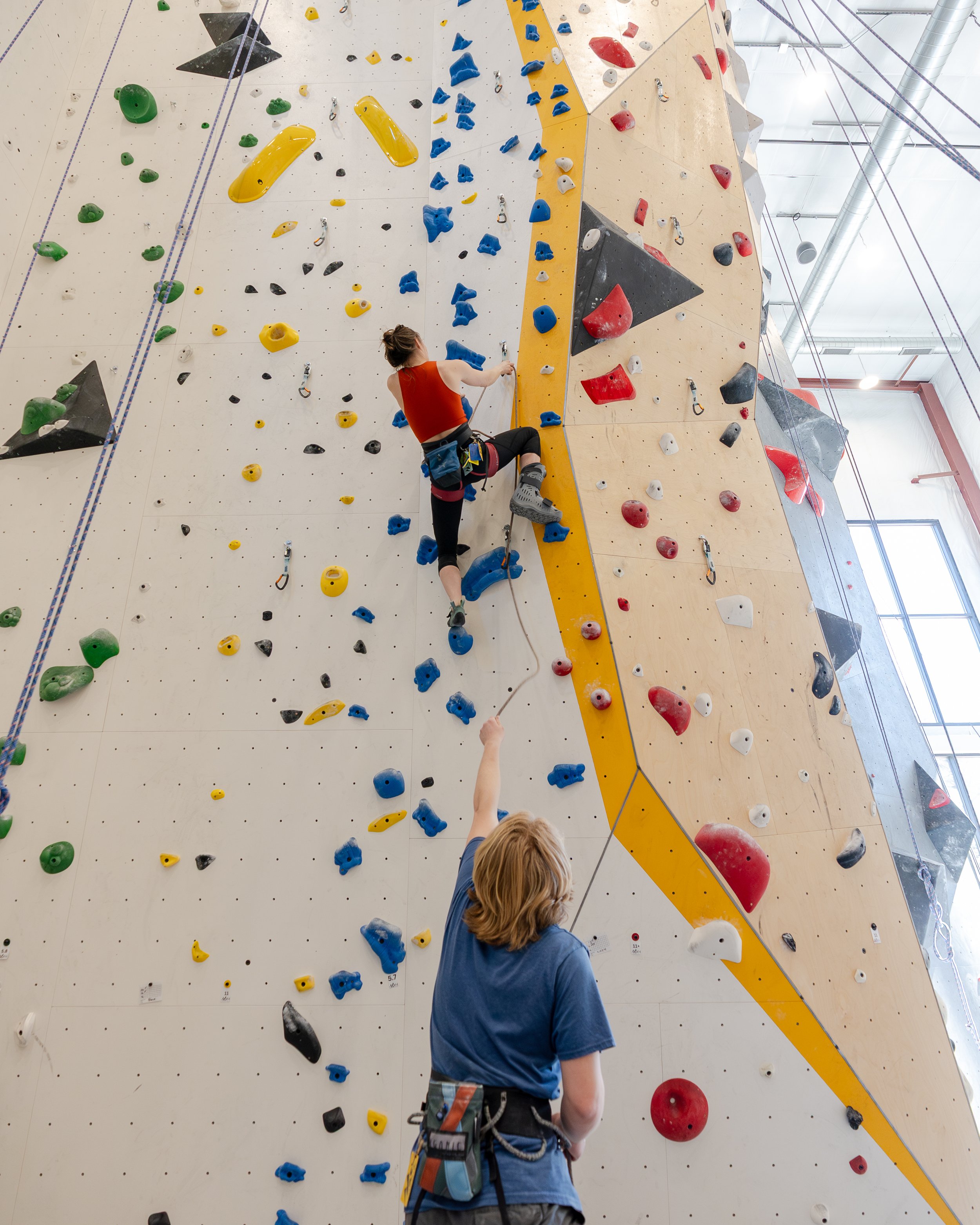 Indoor rock climbing gym with a female climber wearing an orange top and gray climbing shoes ascending a textured wall with various colorful holds, while a belayer in a blue shirt manages the rope.