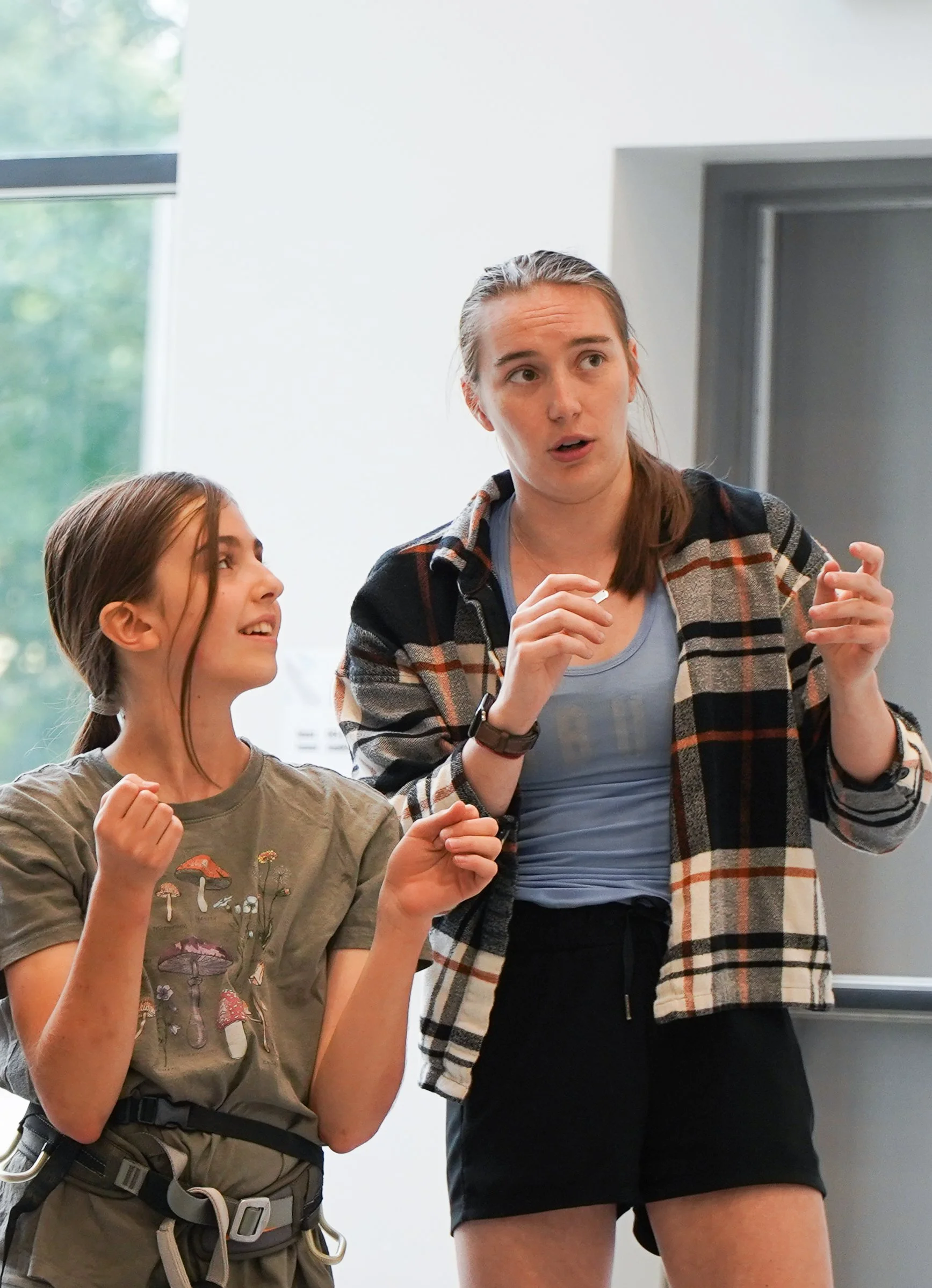 A youth team coach talking with a youth team athlete, with the woman gesturing with her hand and the girl looking up at her. The coach is mirroring moves needed to do the climb the route the youth climber is projecting.