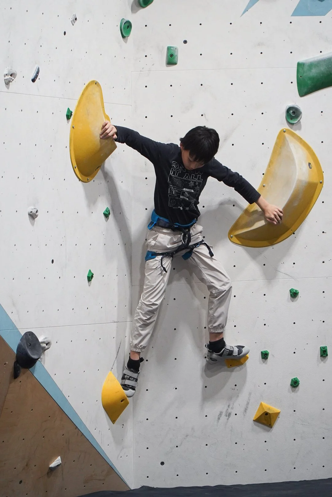 A youth athlete climbing an indoor bouldering wall with yellow and green holds.