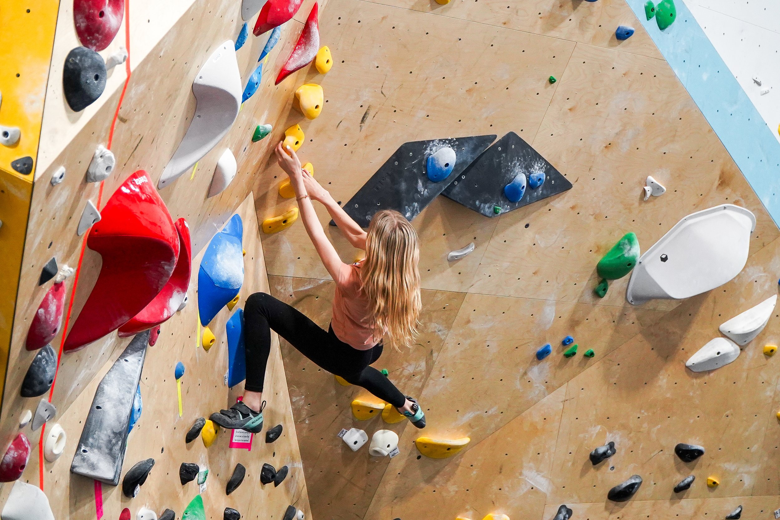 A youth team athlete climbing an indoor bouldering wall with colorful holds. The wall is mostly tan (wood), with streaks of blue and white where the wall angle changes.