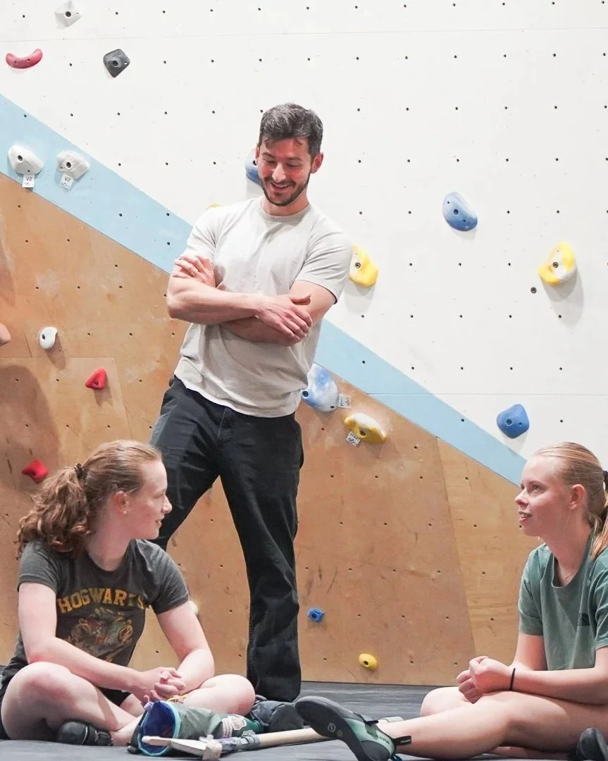 A youth team athlete talking to two youth team athletes who are sitting cross-legged on the floor in an indoor rock climbing gym. The climbing wall behind them is white, tan, and blue with colorful holds.
