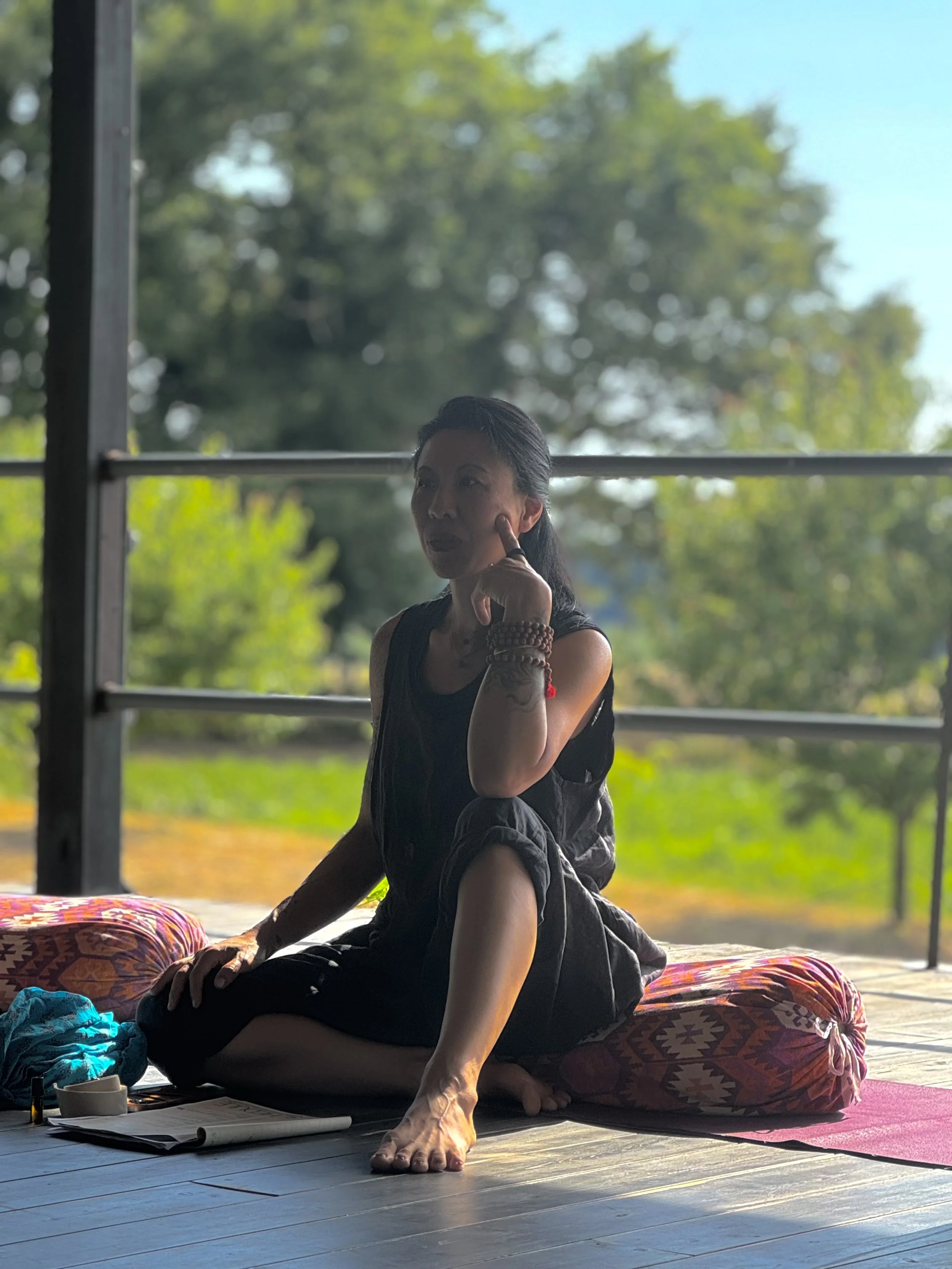 A woman practicing yoga or meditation indoors on a colorful cushion, with open windows showing trees and greenery outside.