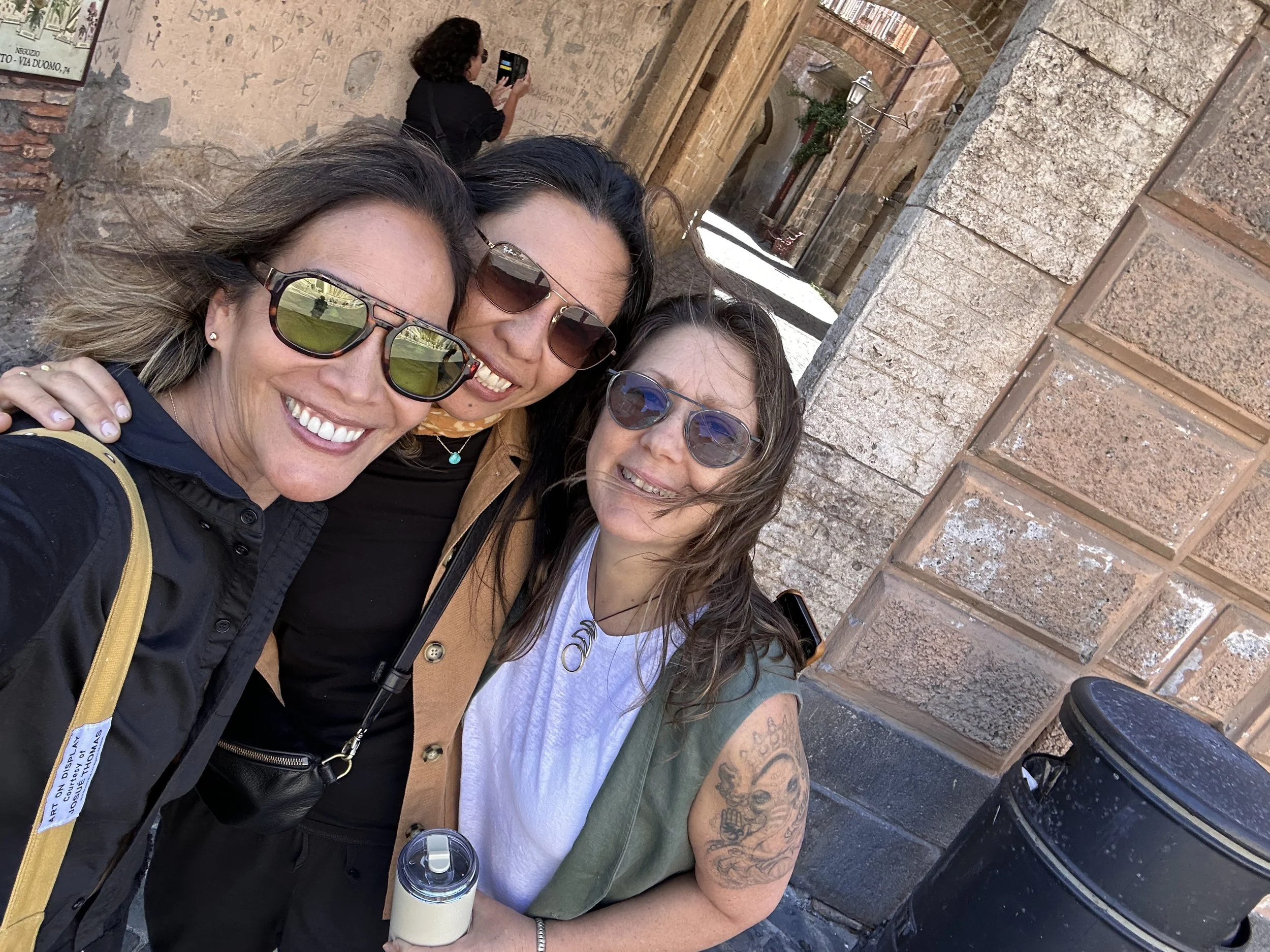 Three women taking a selfie together against a historic brick wall, with one woman in the background using her phone to photograph the scene, and a woman in the foreground smiling with sunglasses.