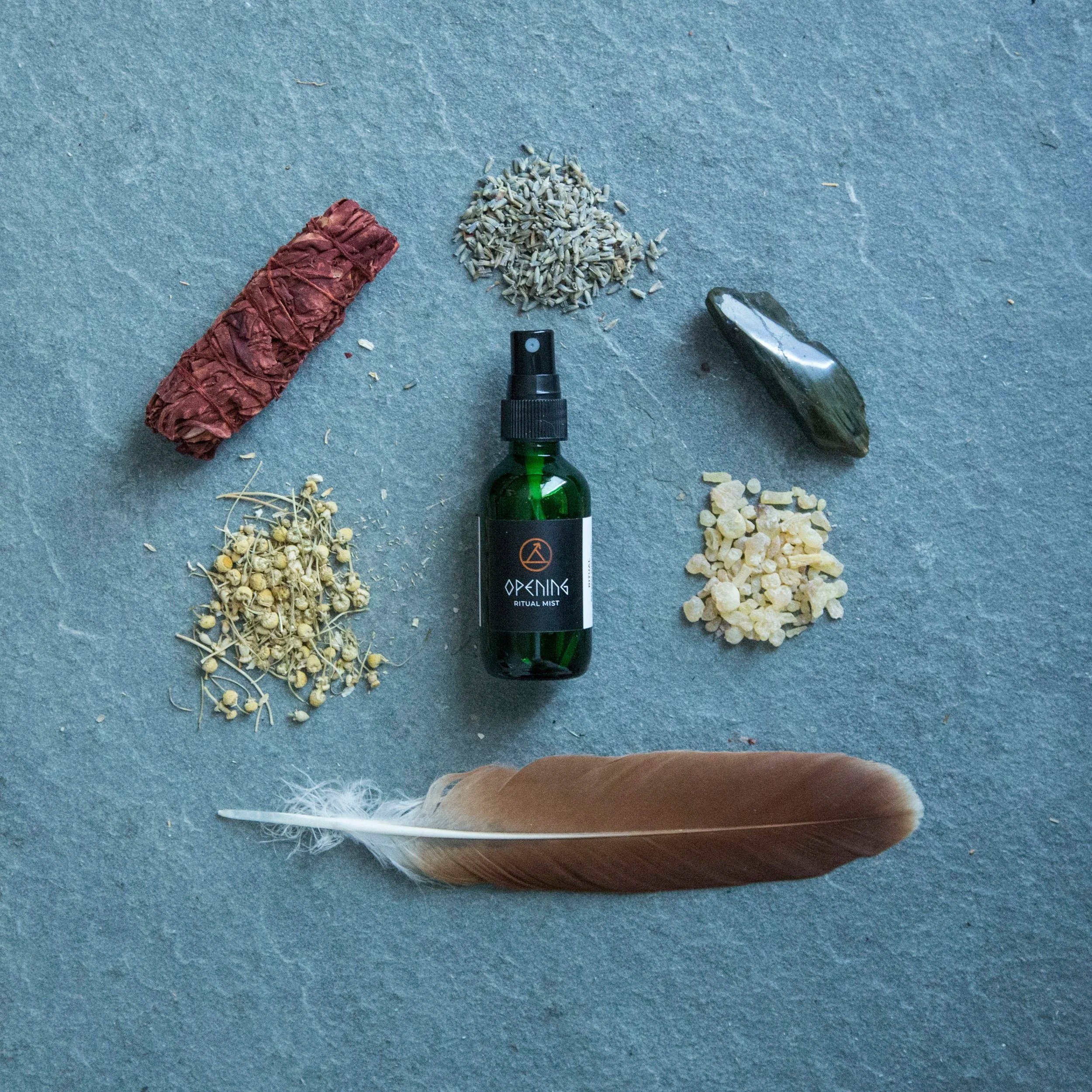 A top-down view of a table with various items including a bundle of herbs wrapped in string, a small pile of dried lavender, a spray bottle labeled 'Opening Ritual Mist,' and scattered dried herbs and flower buds.