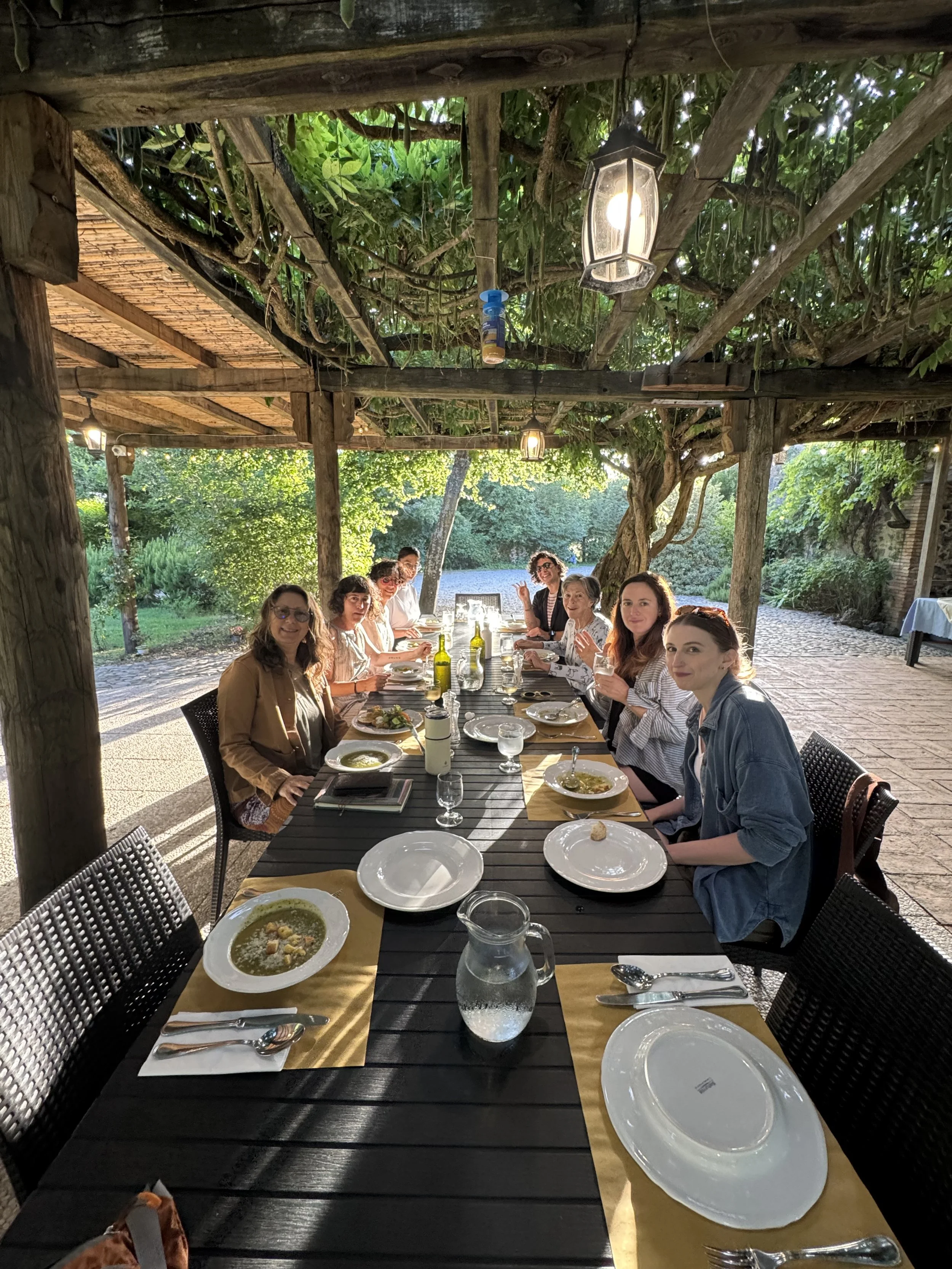 A group of people sitting around a long outdoor dining table under a wooden pergola with greenery. They are having a meal with bowls of soup, plates, glasses, and bottles of wine. The setting is lush and shaded, with sunlight filtering through the le