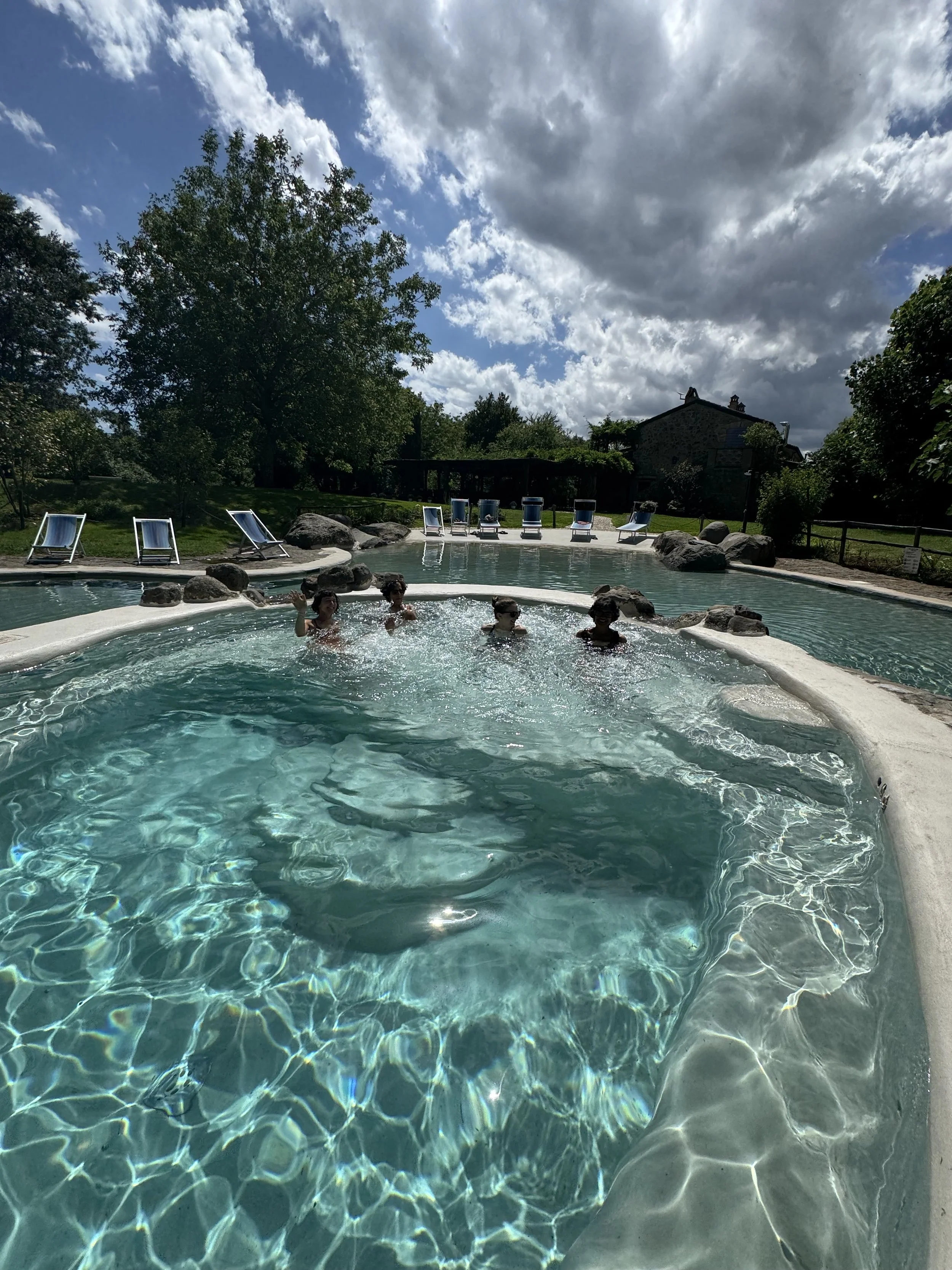 A swimming pool with four people enjoying the water, surrounded by trees and rocks, with lounge chairs at the poolside, and a cloudy sky overhead.