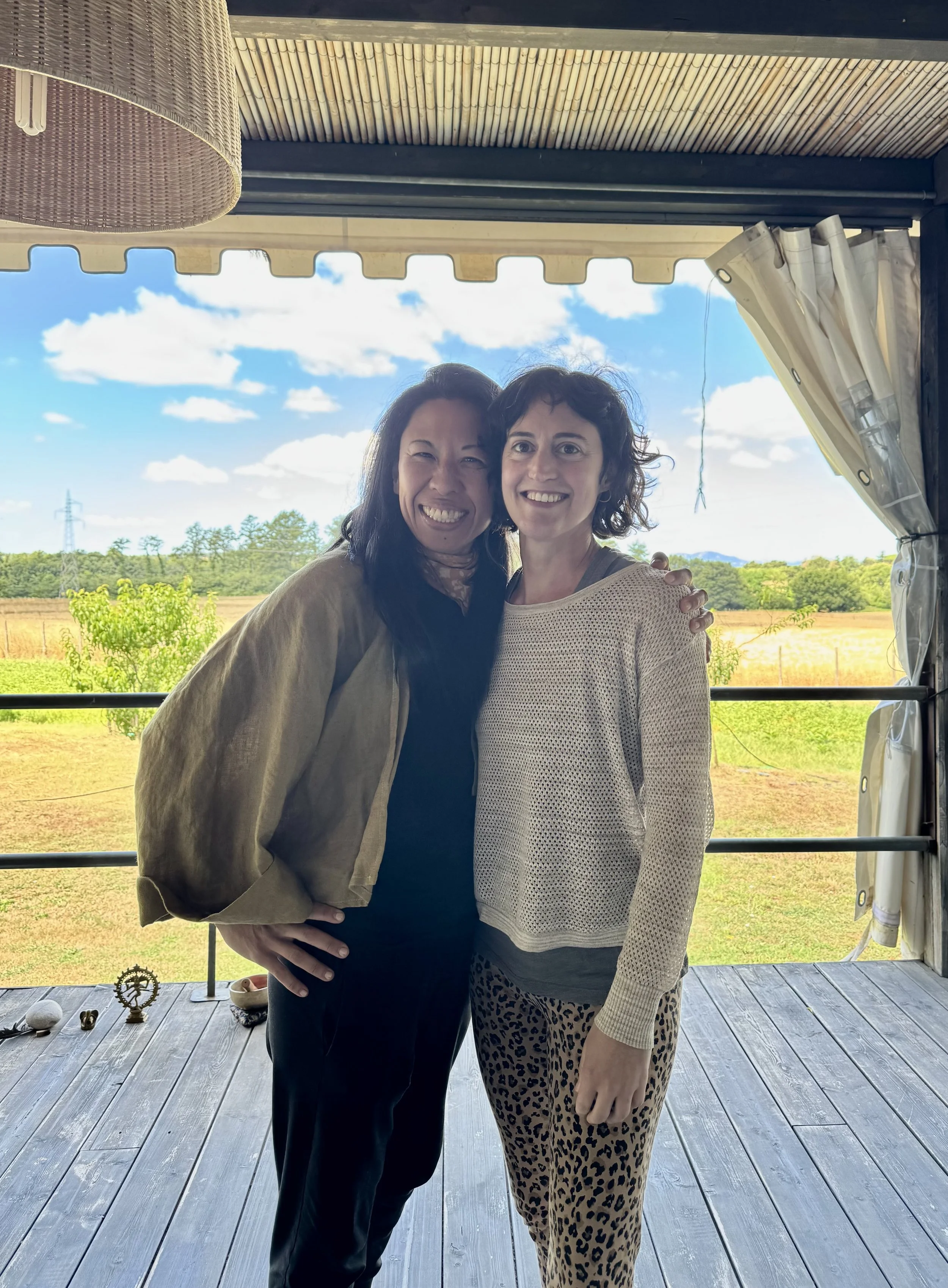 Two women standing close together outdoors, smiling and embracing, with a scenic background of trees and a blue sky with clouds.
